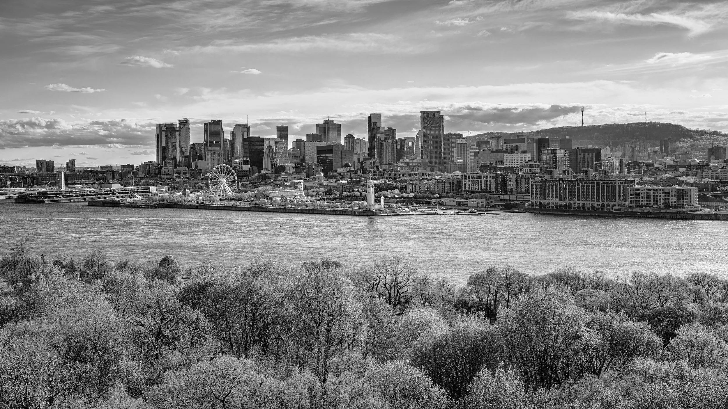 Black and white photo of a city skyline with tall buildings, a ferris wheel, a river in the foreground, and a hill with a tower in the background.