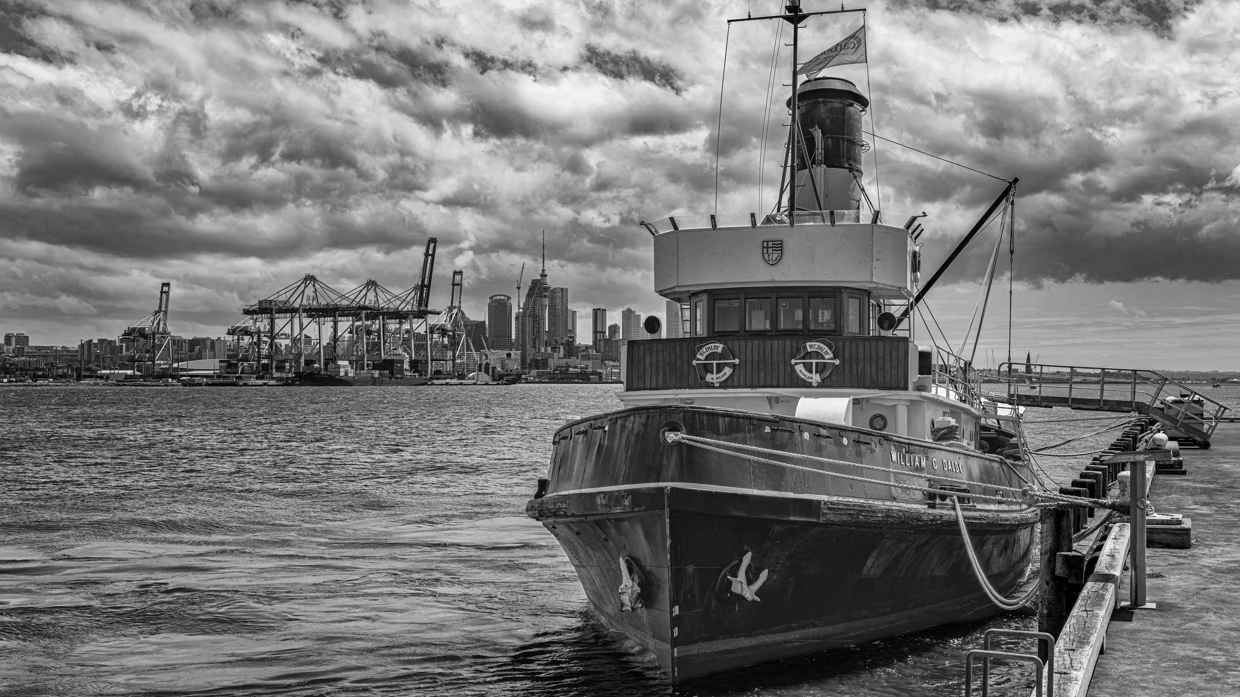 A black and white photo of a boat docked at a pier with a city skyline, including high-rise buildings and cranes, in the background under cloudy skies.