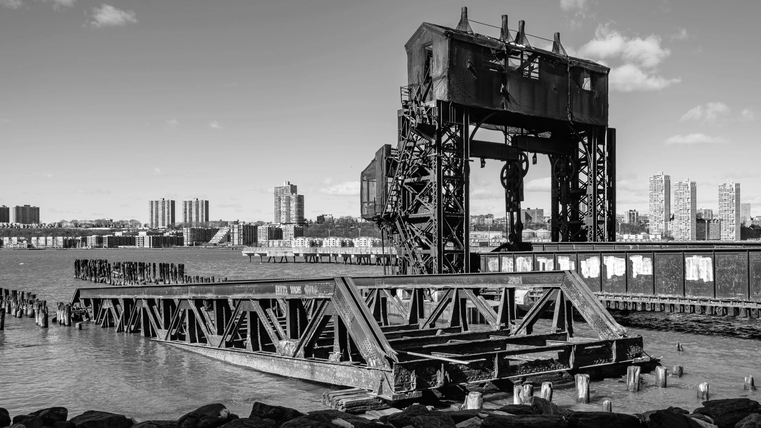 Black and white photo of an old, dilapidated waterfront structure with rusted metal, wooden steps, and scaffolding, overlooking a city skyline with high-rise buildings across a body of water. Manhattan, New York, USA.