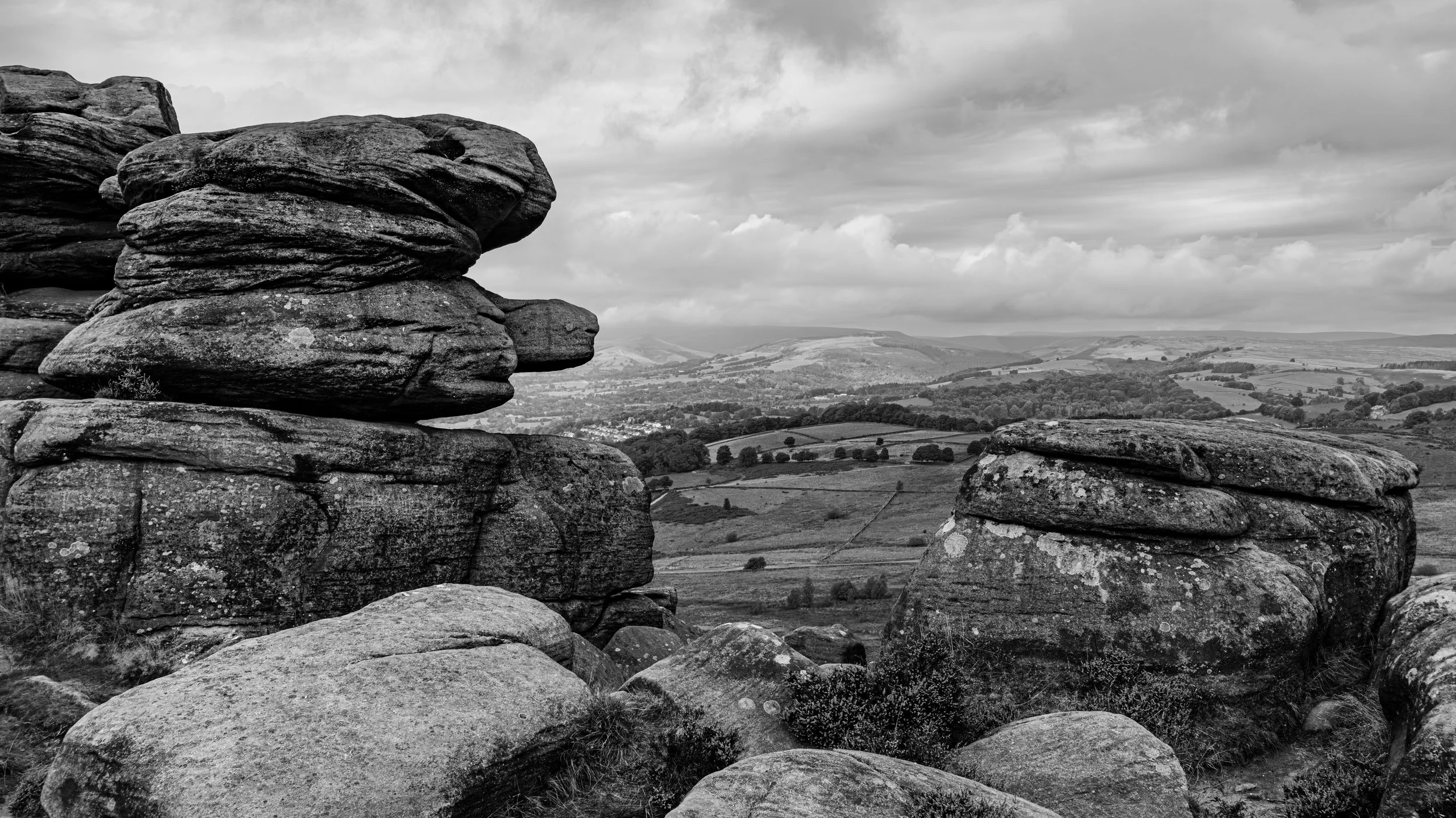 Black and white landscape view with large weathered rocks in the foreground and rolling hills, trees, and cloudy sky in the background.