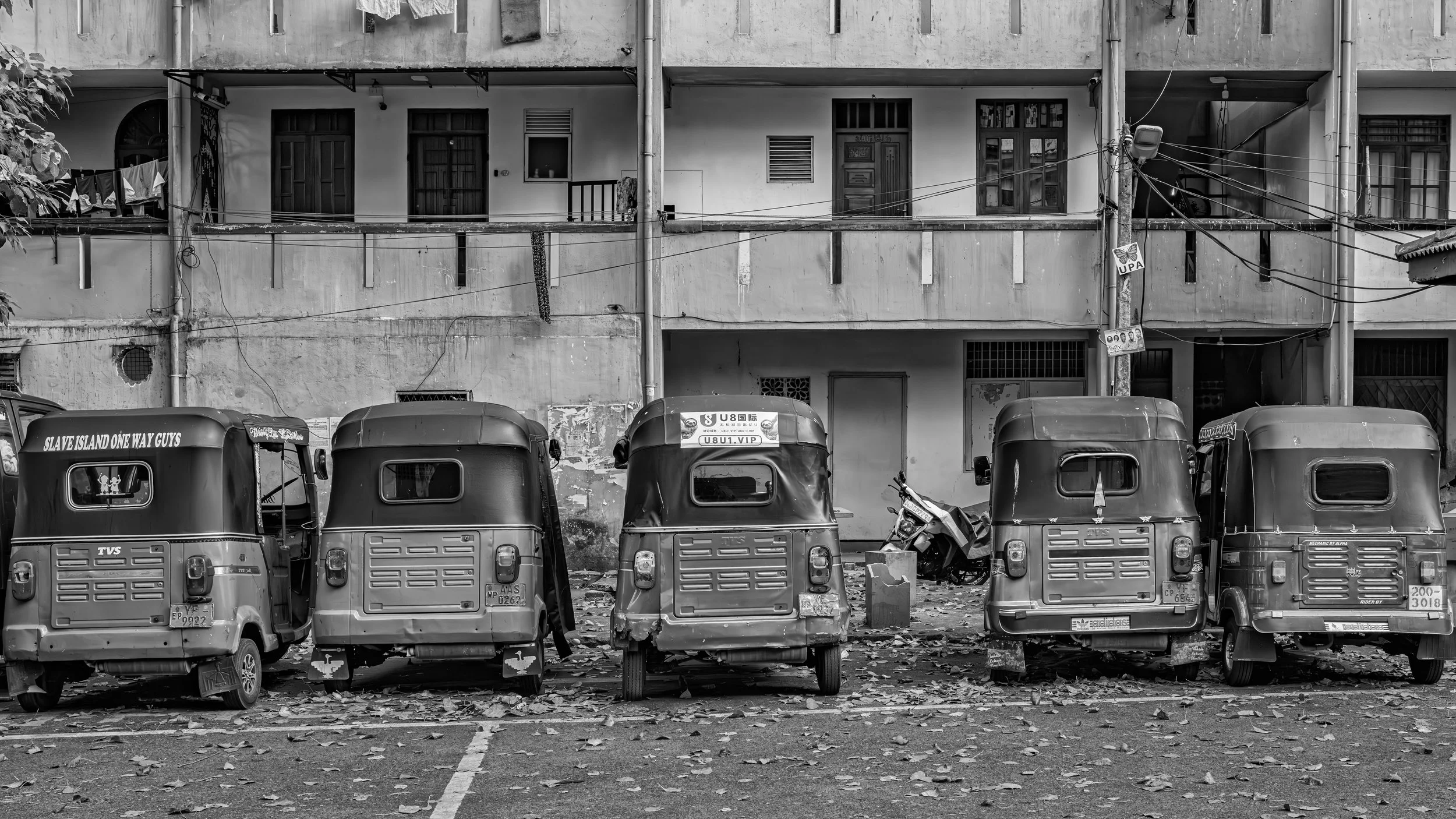 Black and white photo of a parking lot with four auto rickshaws and a motorcycle in front. Behind them is a building with multiple apartments, visible windows, doors, and electrical wires across the scene. Fallen leaves are scattered on the ground.
