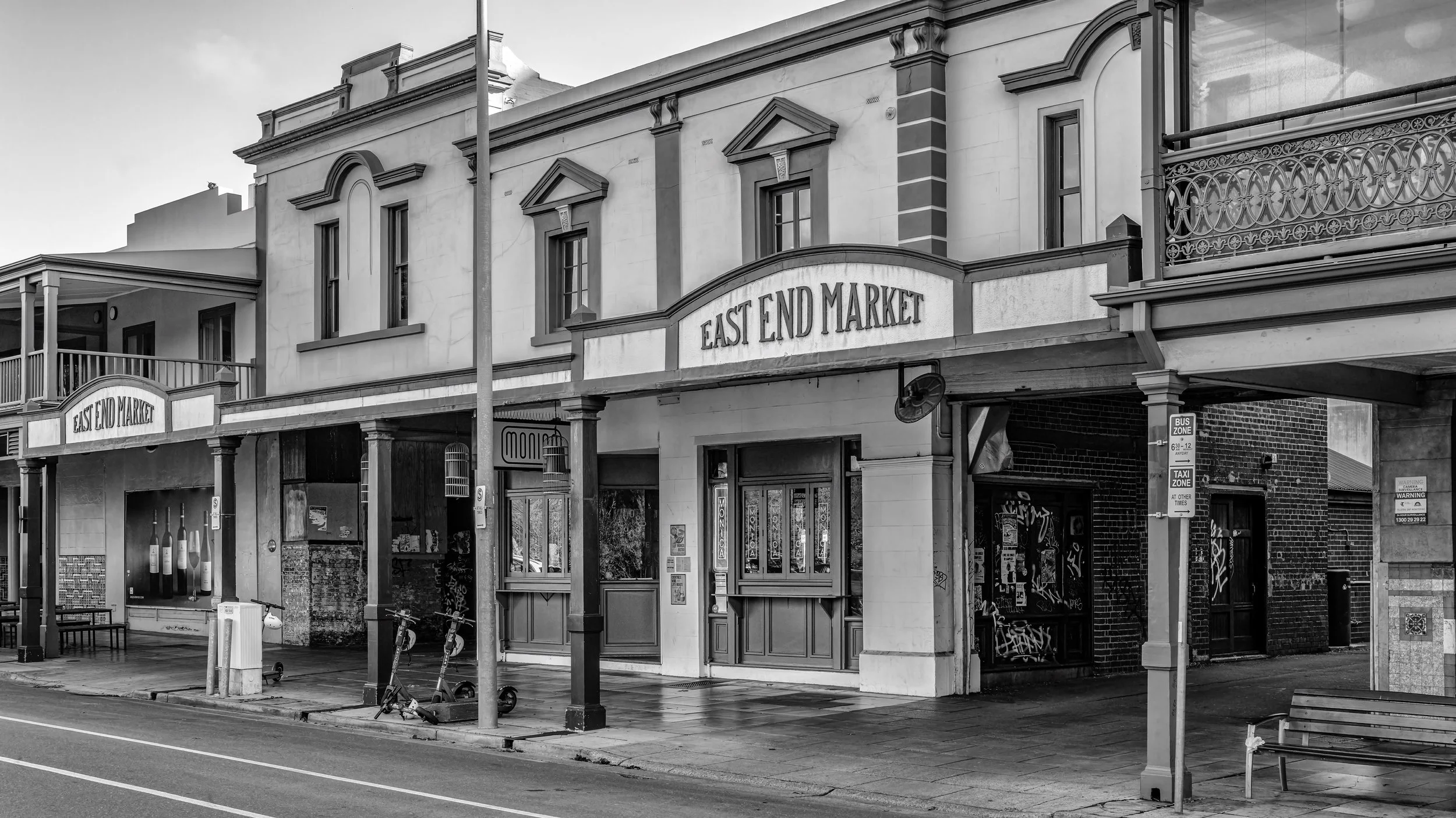 Black and white photo of a storefront with a sign that reads 'East End Market'. The building has decorative windows and a balcony with ornate railing. There are bicycles and a bus zone sign in the foreground. Adelaide, Australia.