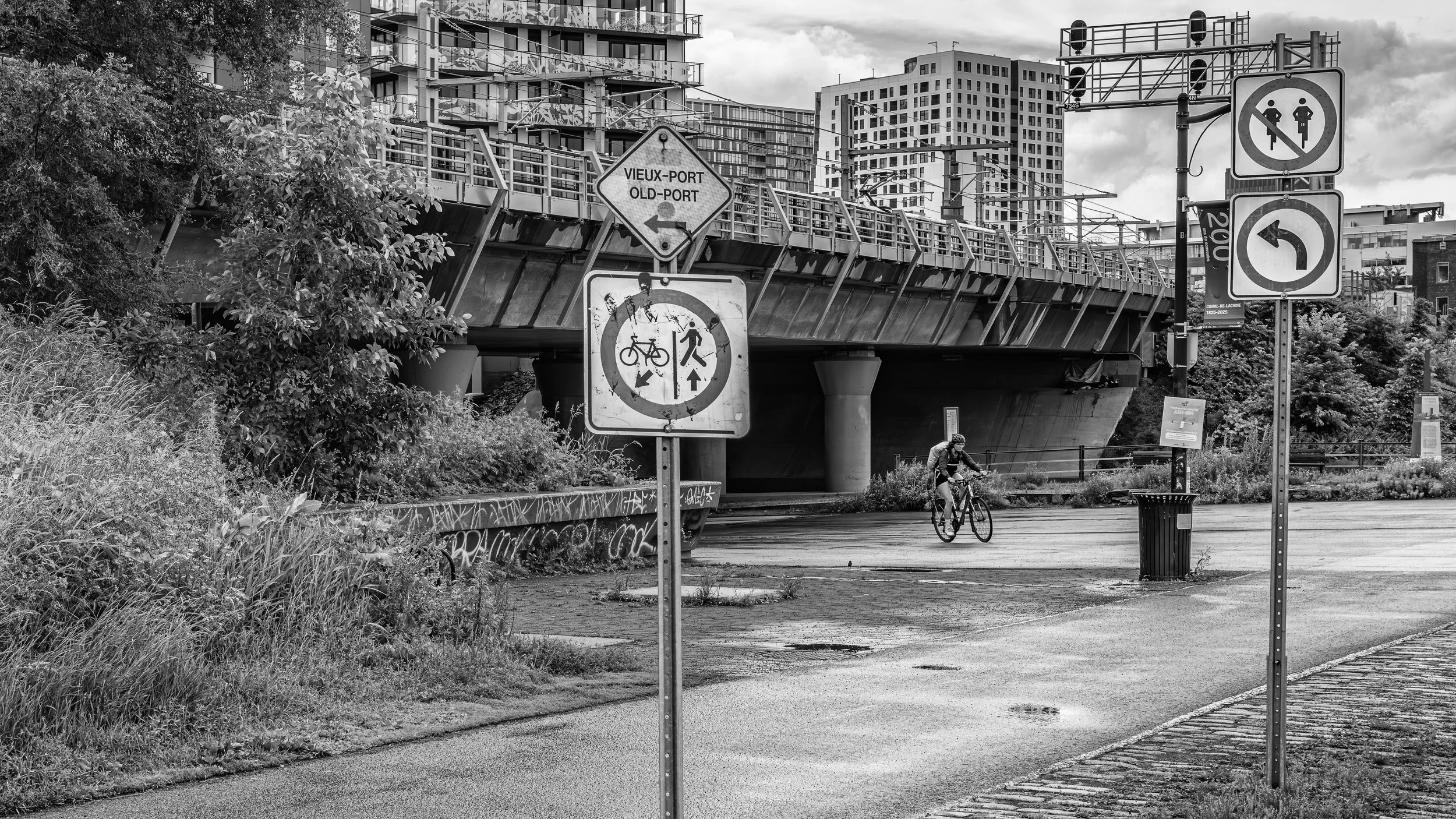 A city scene in black and white showing a cyclist riding on a path under a bridge. There are multiple signs, including one indicating a bike and pedestrian zone and another indicating a left turn. Tall buildings are visible in the background, and the