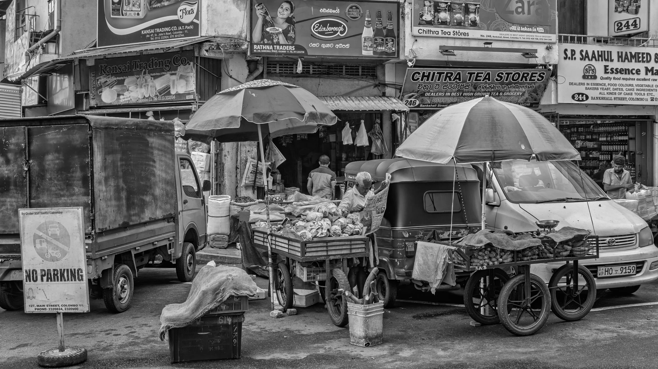 Street market scene with vendors selling goods under umbrellas, cars parked nearby, and various shop signs in the background.