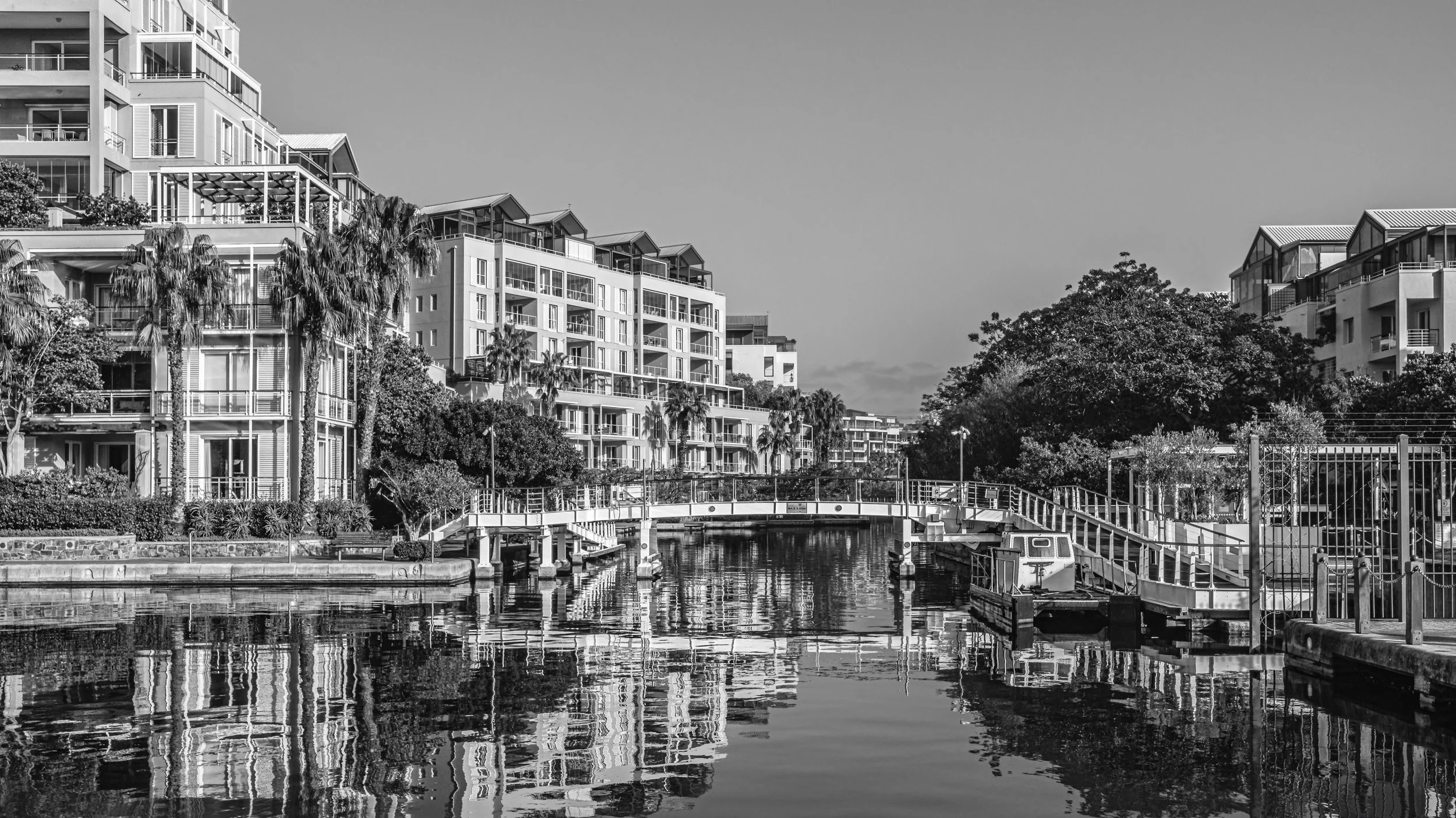 Modern residential buildings along a waterway with small boats, trees, and a pedestrian bridge, in black and white. Cape Town, South Africa.