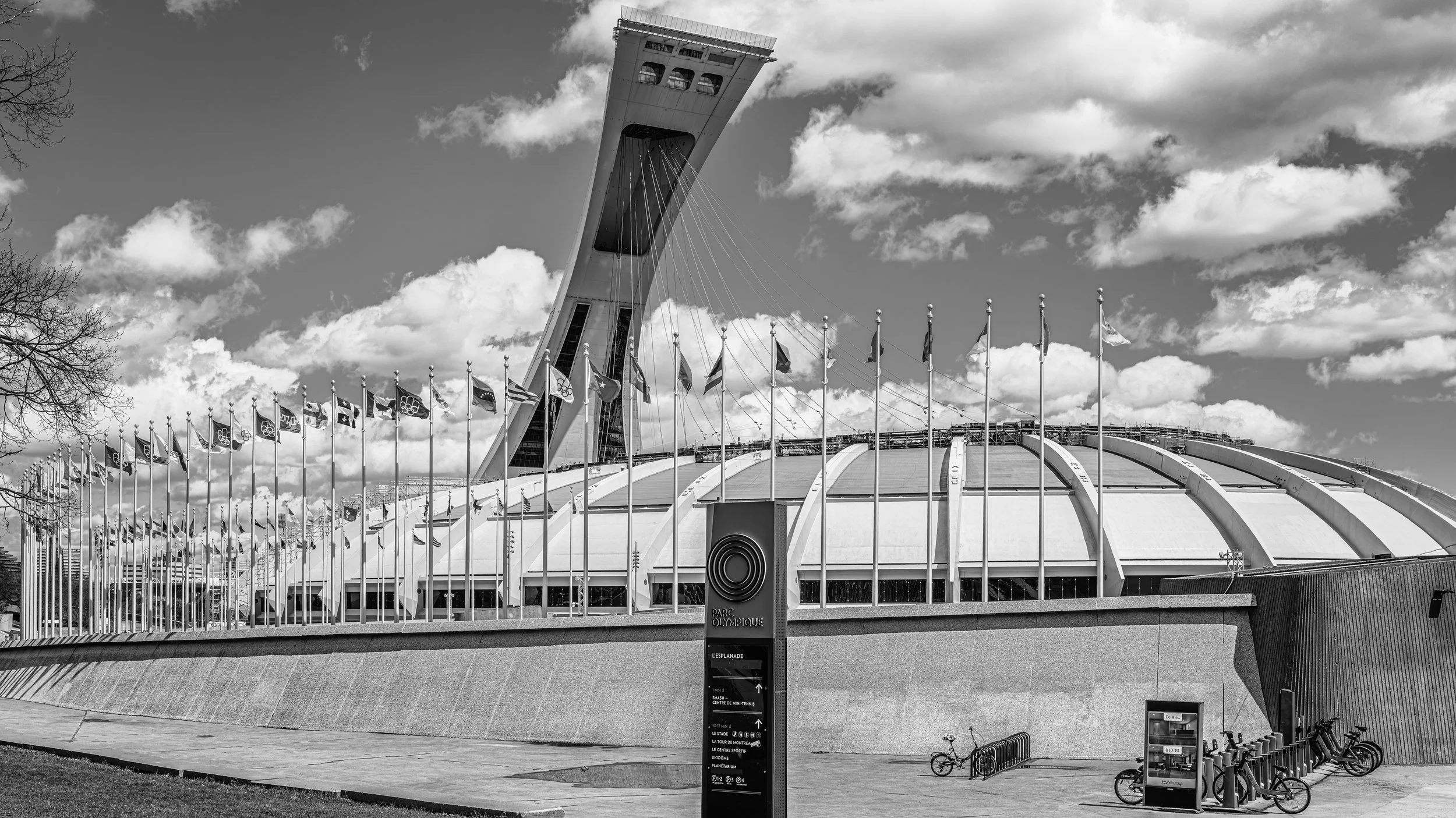 Black and white photo of Paris Olympic Stadium with a tall, angular tower and a curved roof. Multiple flags are displayed along a fence surrounding the stadium. Traffic signs and bicycles are visible in the foreground, with a cloudy sky overhead.