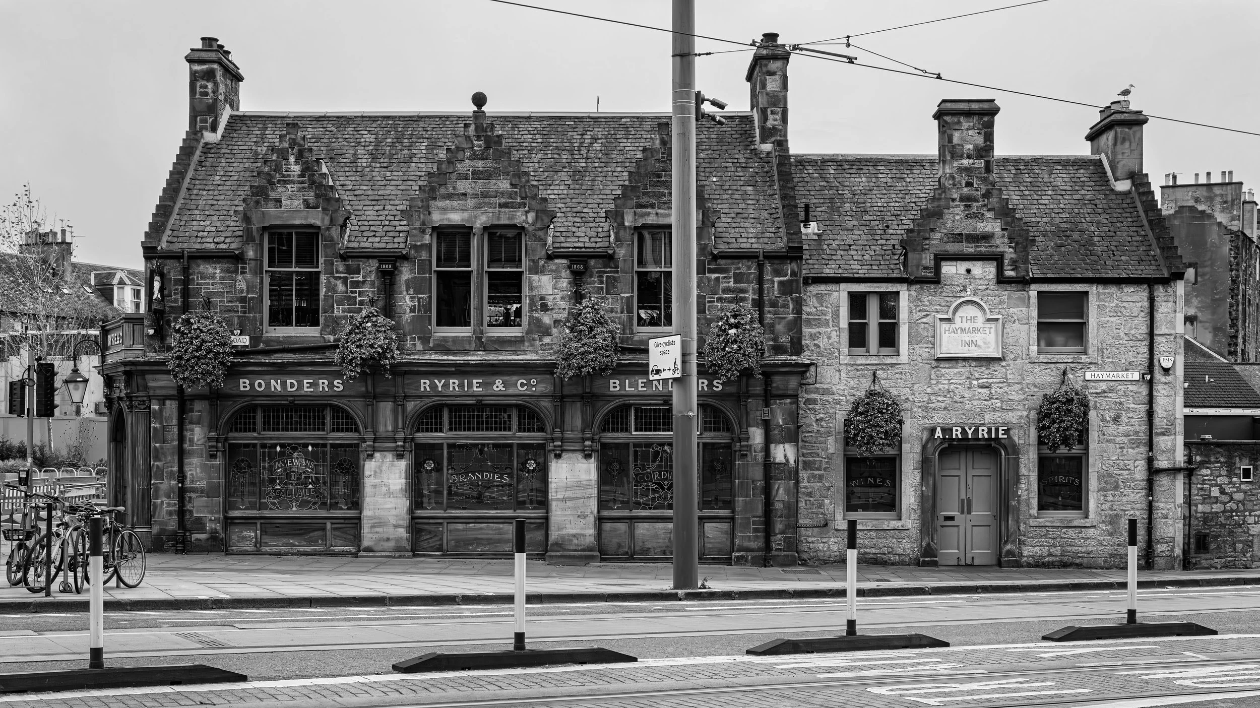 Black and white photo of an old stone building with a sign reading 'The Haymarket Inn' and storefront labels for 'Bonders Ryrie & Co. Blenders,' with bicycles and a street scene in front.