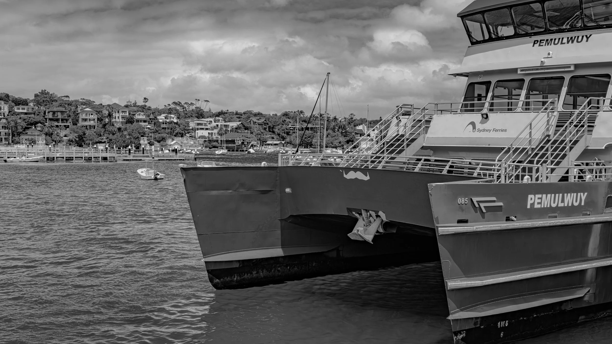 Black and white photo of a ferry boat named PEMULWUY docked in the water with a residential hillside village in the background.