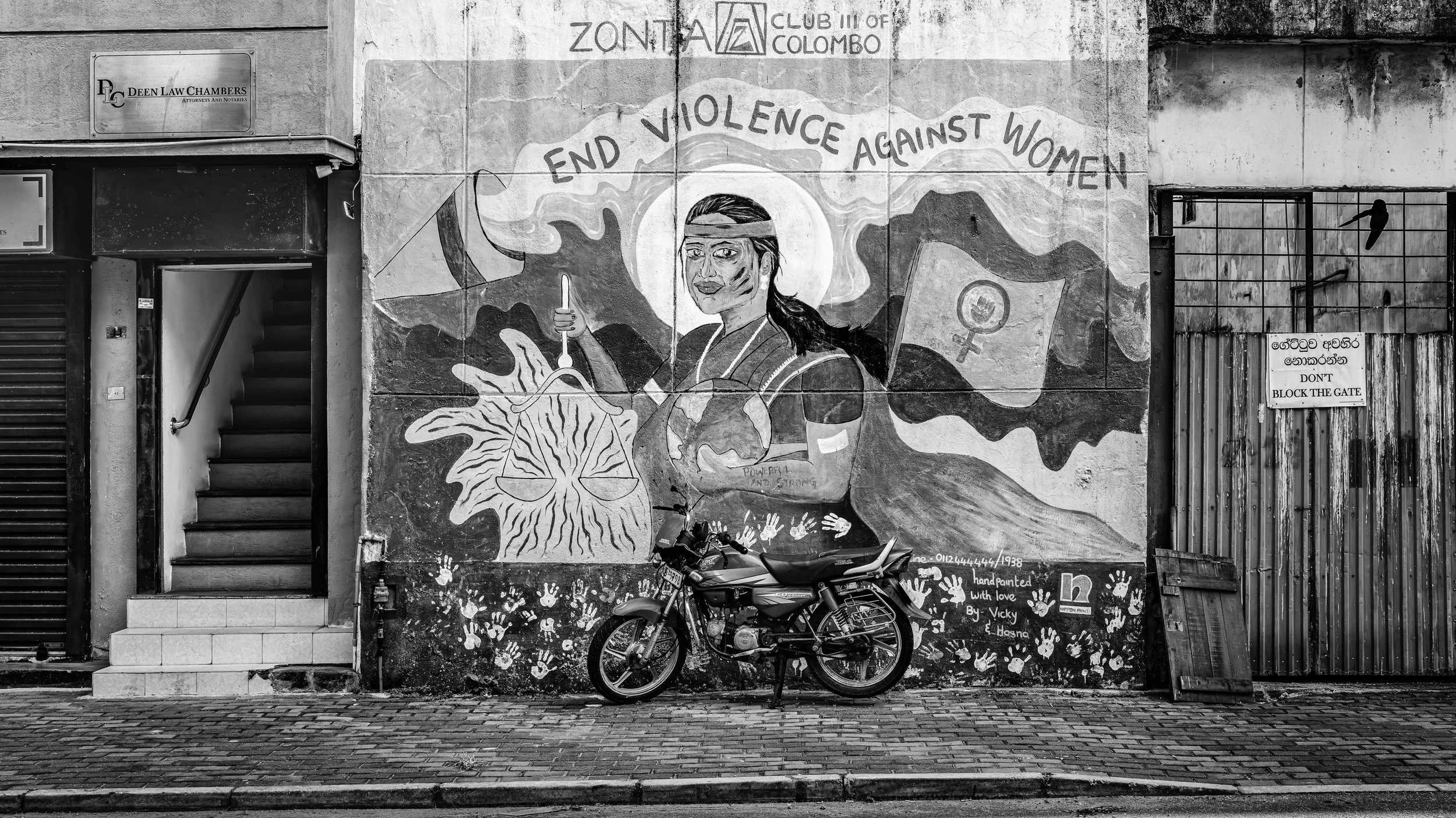 A black-and-white mural on an urban wall protesting violence against women, featuring a woman holding a candle, scales of justice, and a globe. Text above reads 'End Violence Against Women.' A parked motorcycle is in front of the mural, and a sign on