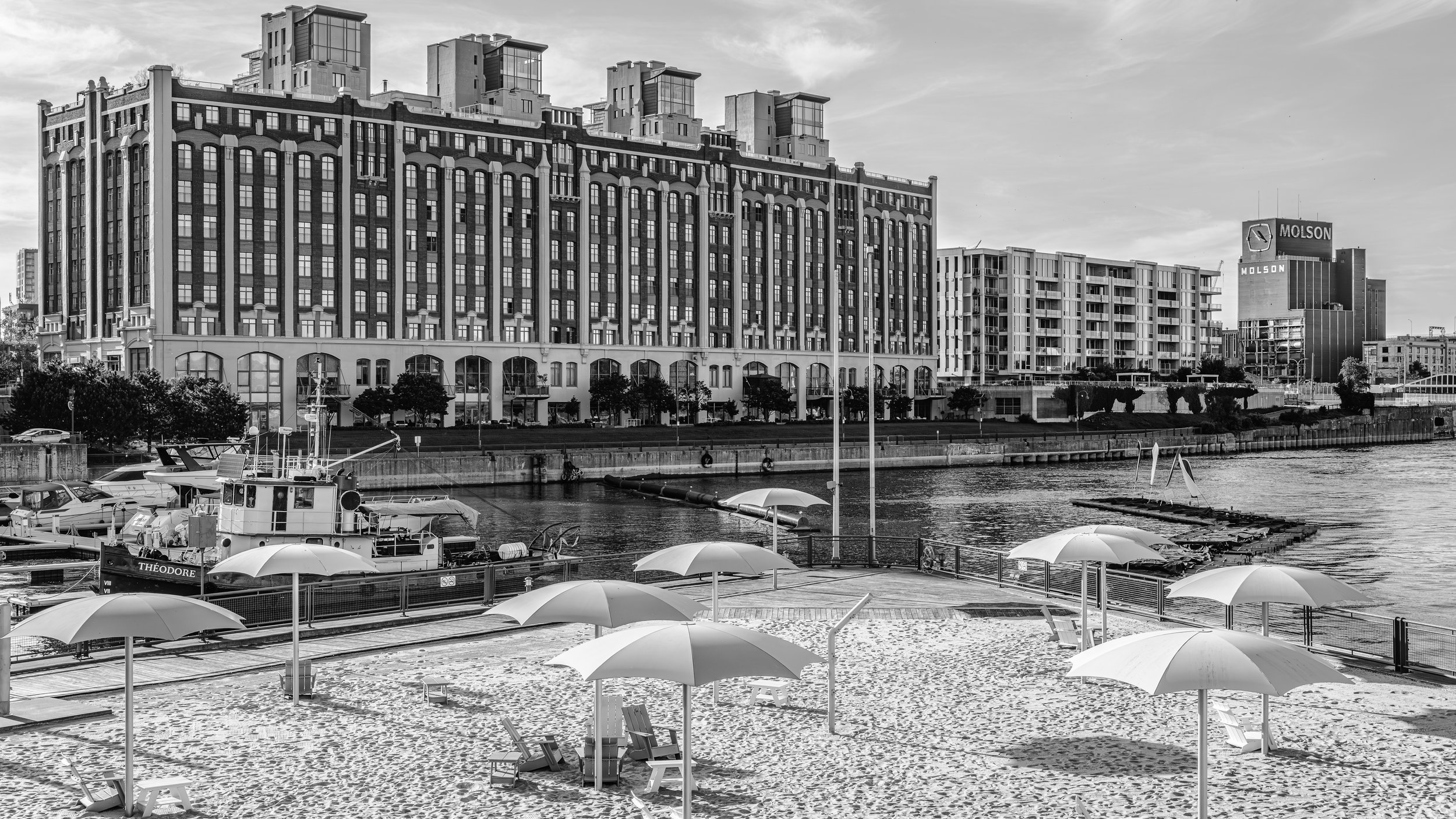 Black and white photo of a city skyline with tall buildings and a river in the foreground. There is a marina with boats and umbrellas at a waterfront park.