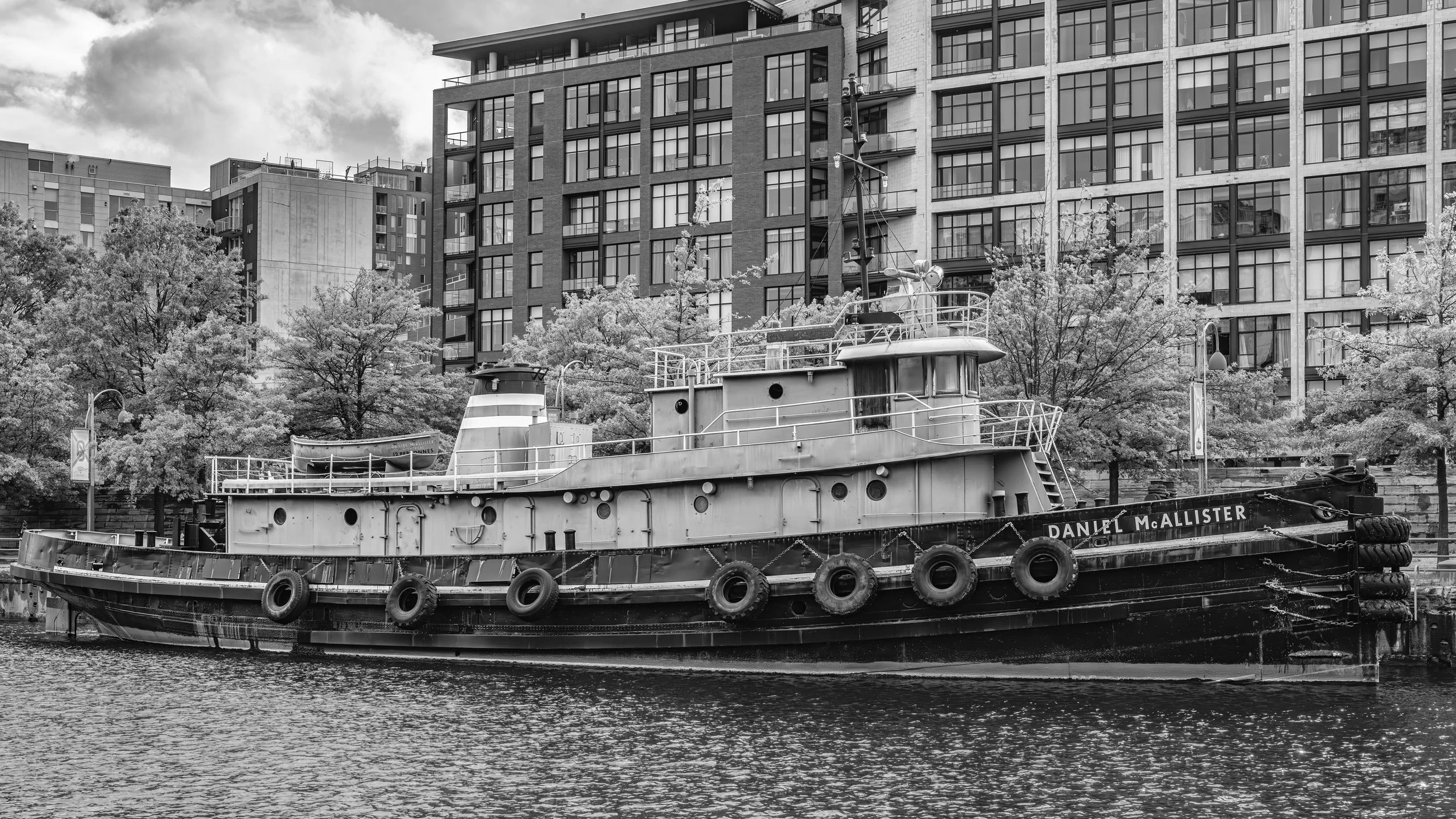 Black and white photo of an old boat named 'Daniel McAllister' docked along a waterway, with modern apartment buildings and trees in the background.