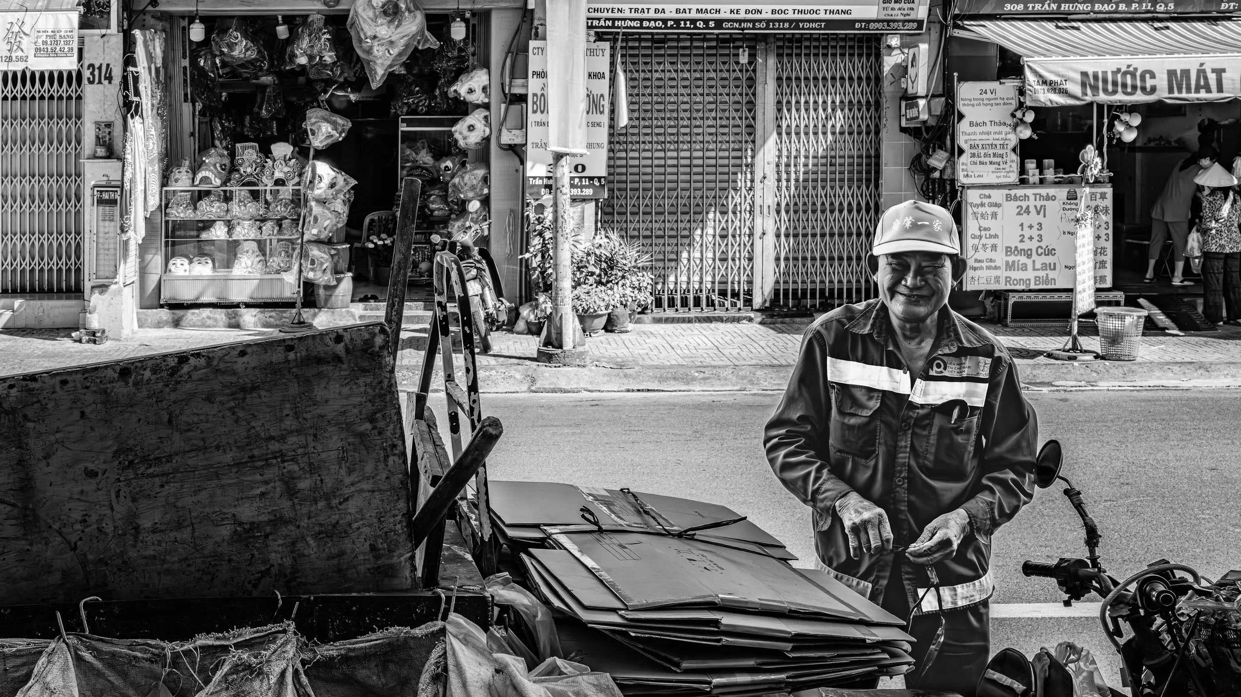 A street scene in black and white showing a man with a cap and smiling face standing beside a bicycle and boxes. In the background, there are shopfronts with closed metal shutters, signs in Vietnamese, and various street goods like masks and plastic 