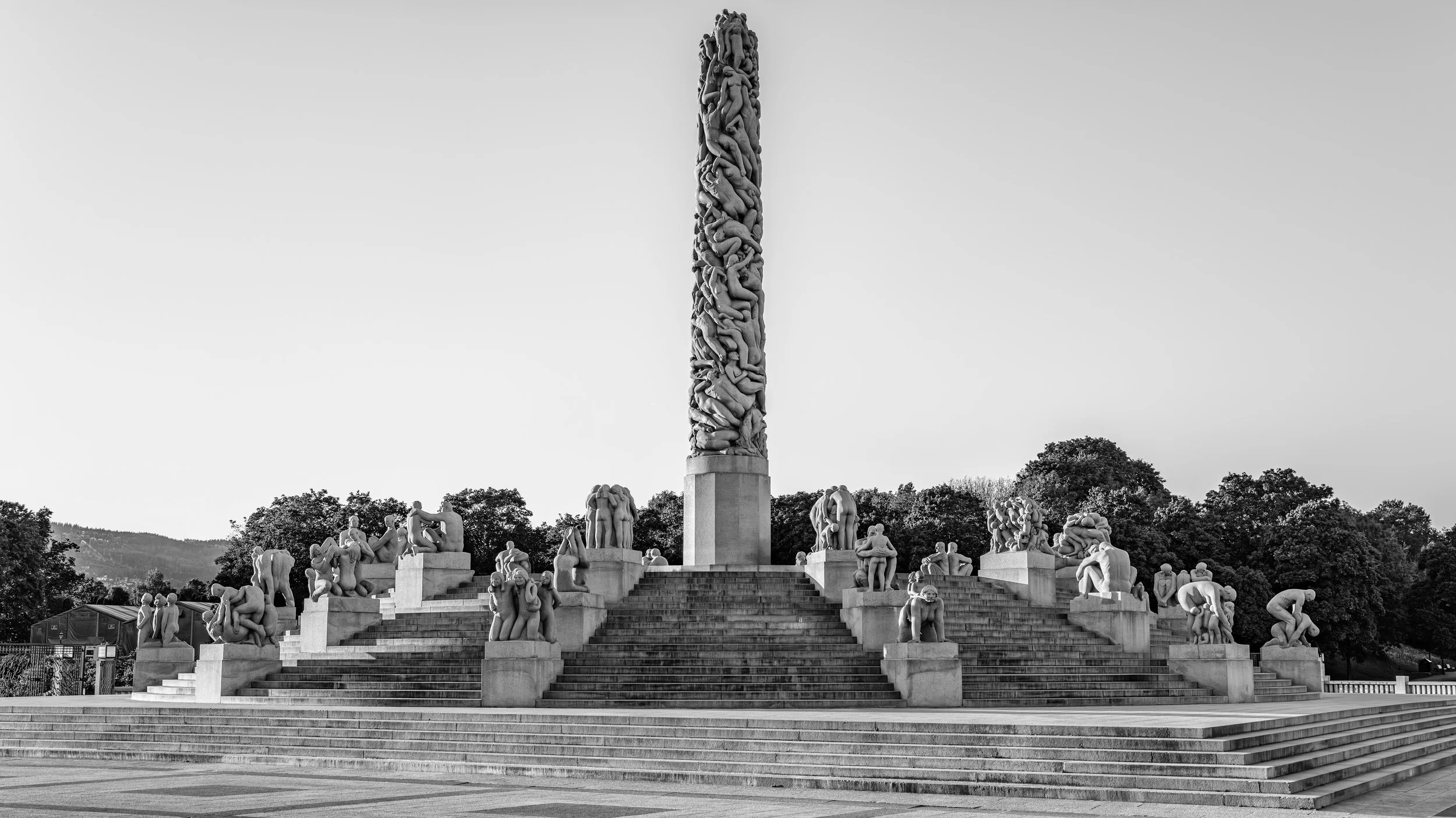 A large outdoor monument featuring numerous human figures carved in stone, with a tall column of intertwined bodies at the center. The monument is set on a broad staircase surrounded by trees, with a clear sky overhead.