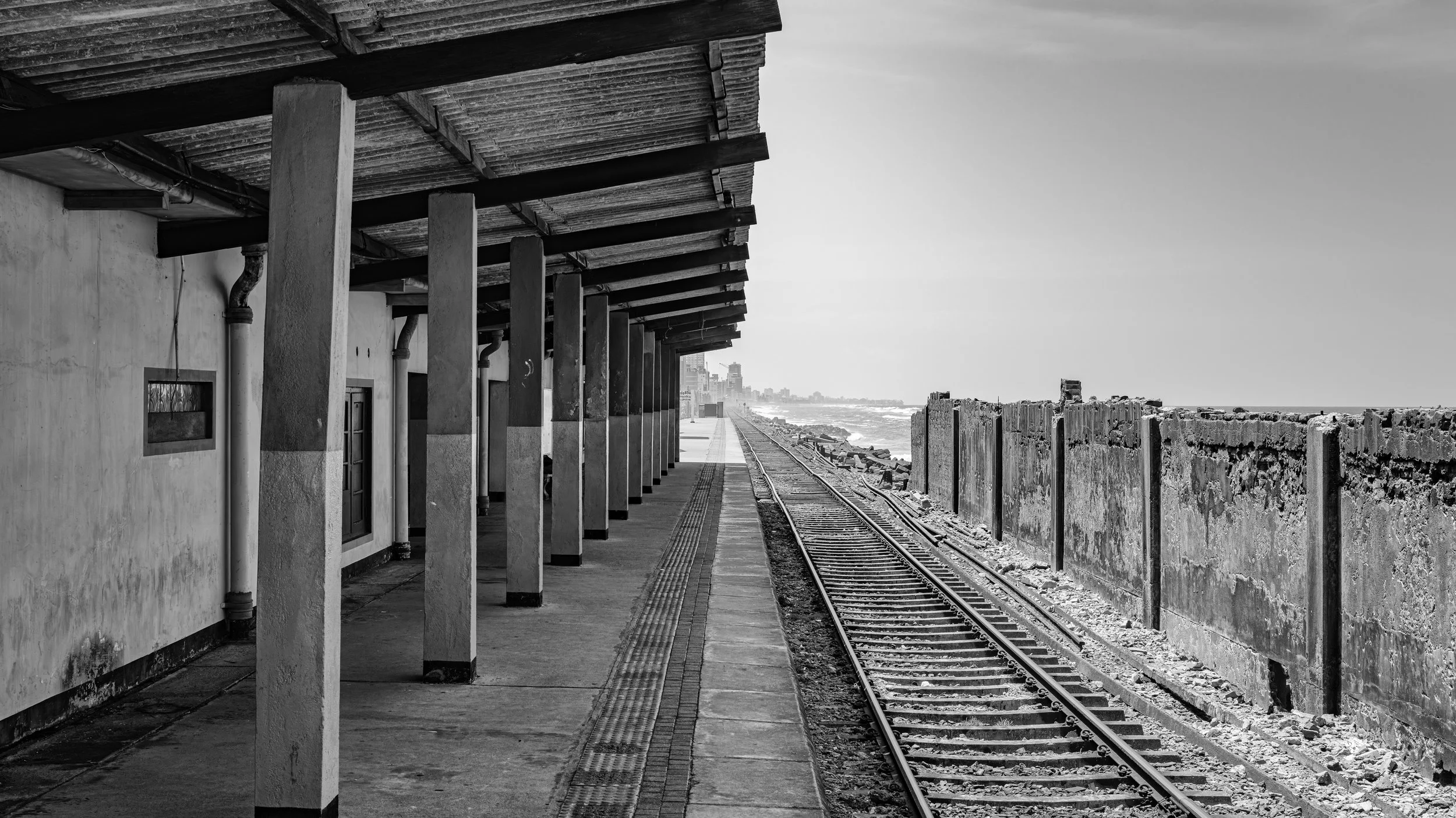 Black and white photo of a seaside train platform with a building on the left, train tracks running along the platform, and a crashing ocean in the background.