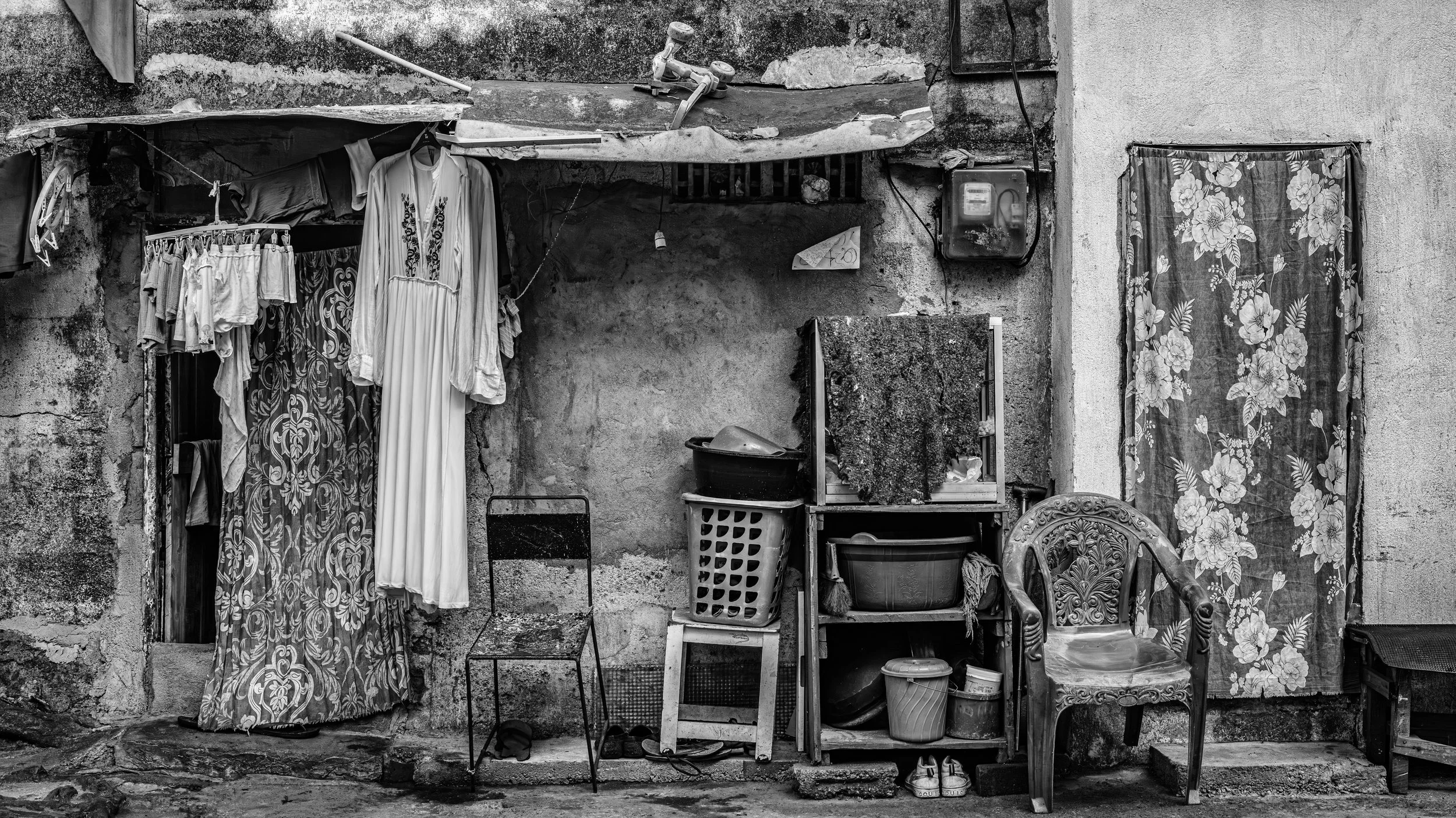 A black and white photo of a worn, textured outdoor wall with various household items, including clothing hanging on a line, a plastic laundry basket, chairs, and a curtain with a floral pattern.