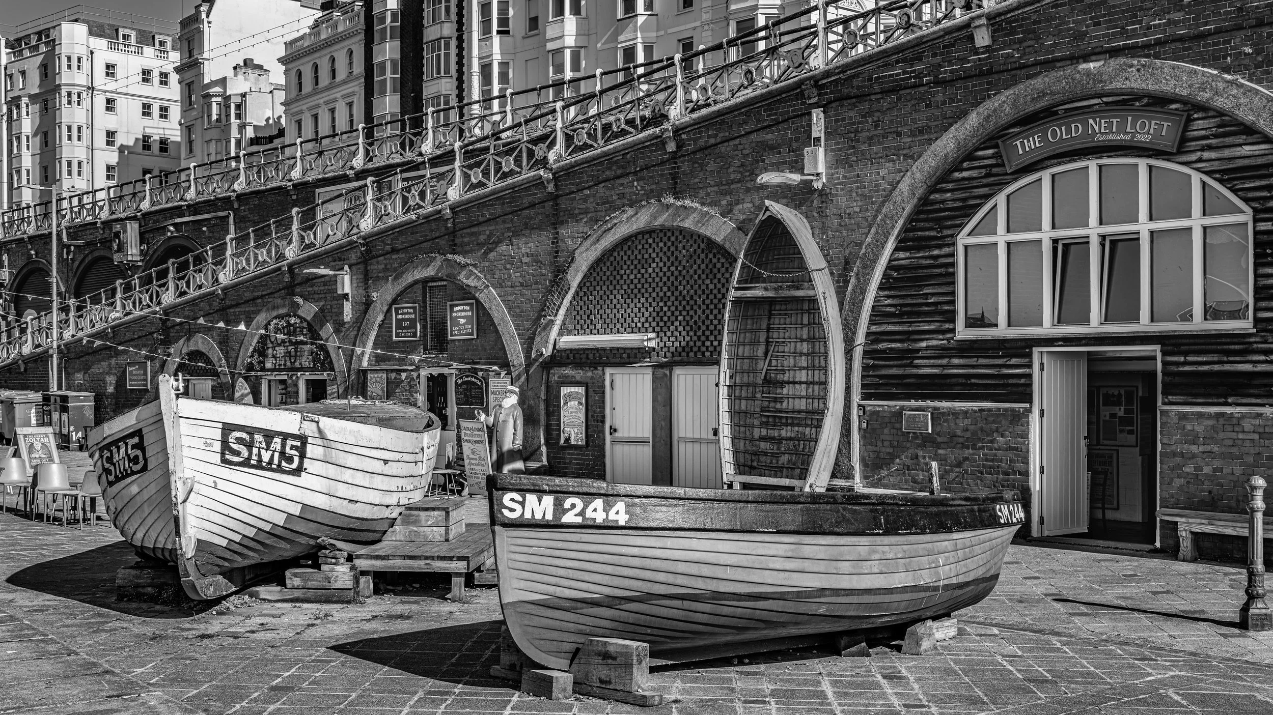 Two docked boats with registration numbers SM5 and SM244 are positioned in front of a brick building with arched windows and a sign that reads 'The Old Net Loft.' The scene is in a maritime or harbor area with chairs, signs, and a person walking near