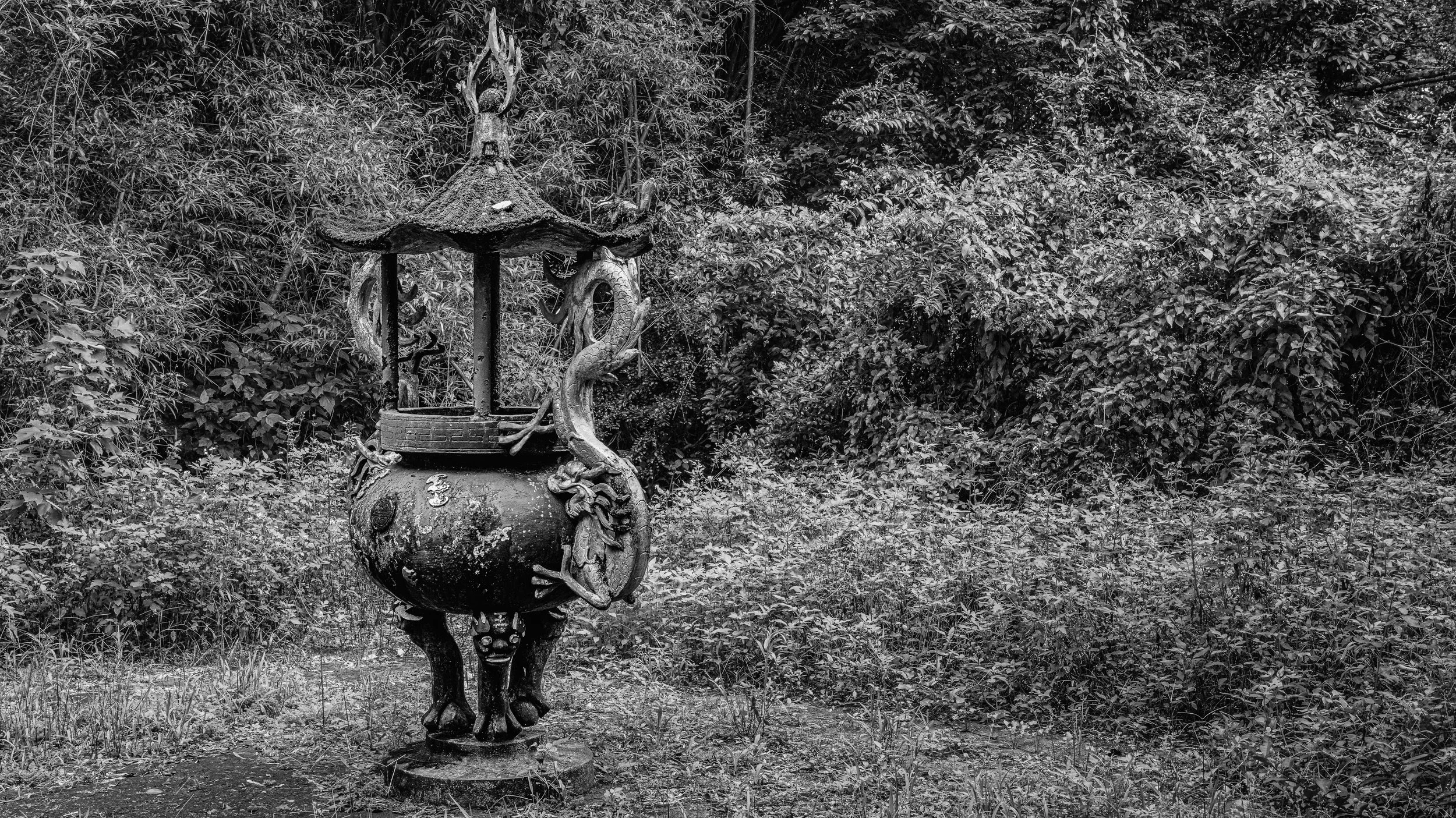 An ornate metal lantern with dragon sculptures wrapped around its top and sides, set against a dense background of leafy bushes.