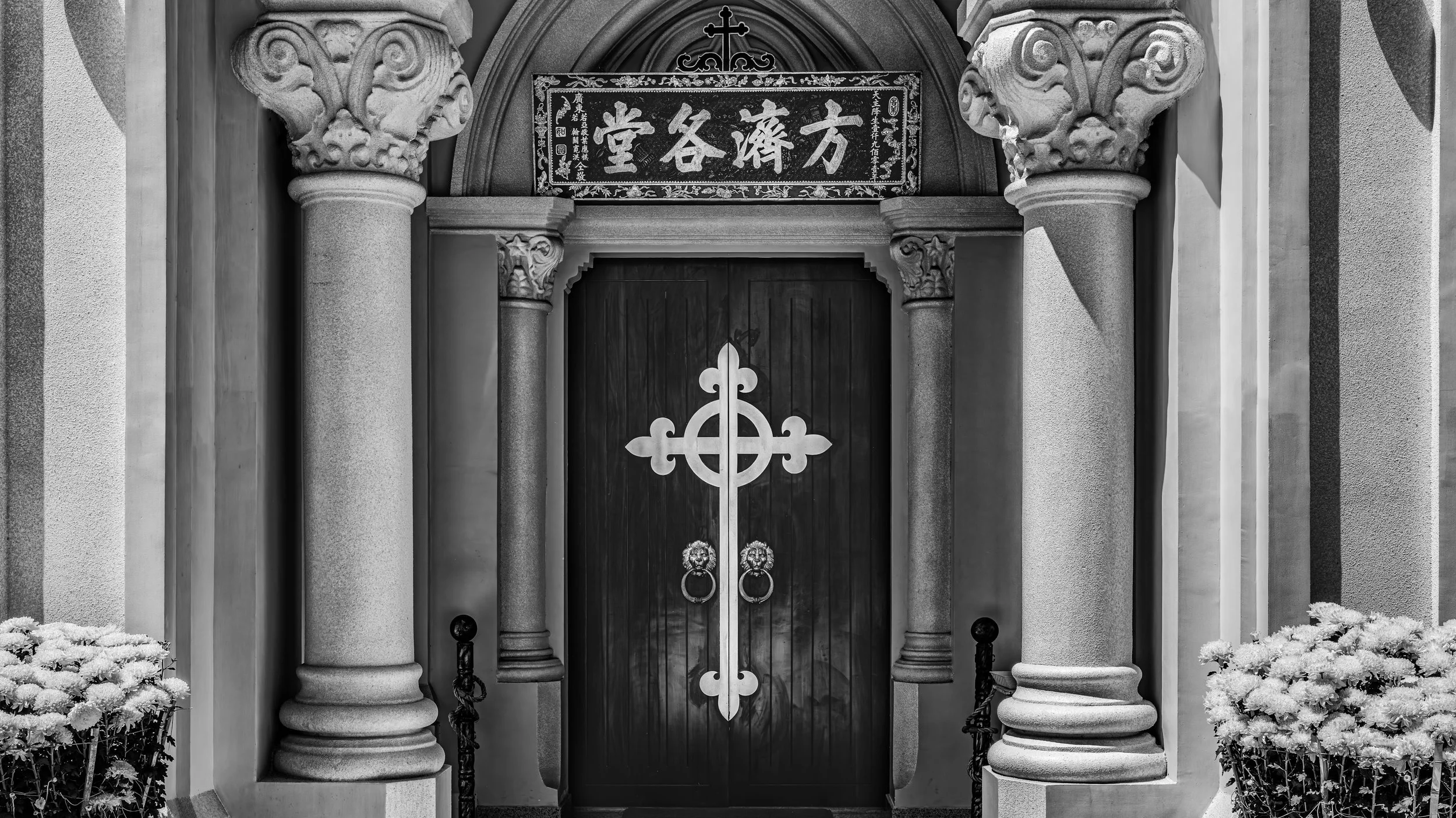 A black and white photograph of a church entrance with two tall stone columns on each side of a wooden door. The door has a decorative iron design and two lion head door knockers. Above the door, there is a sign in Chinese characters surrounded by in