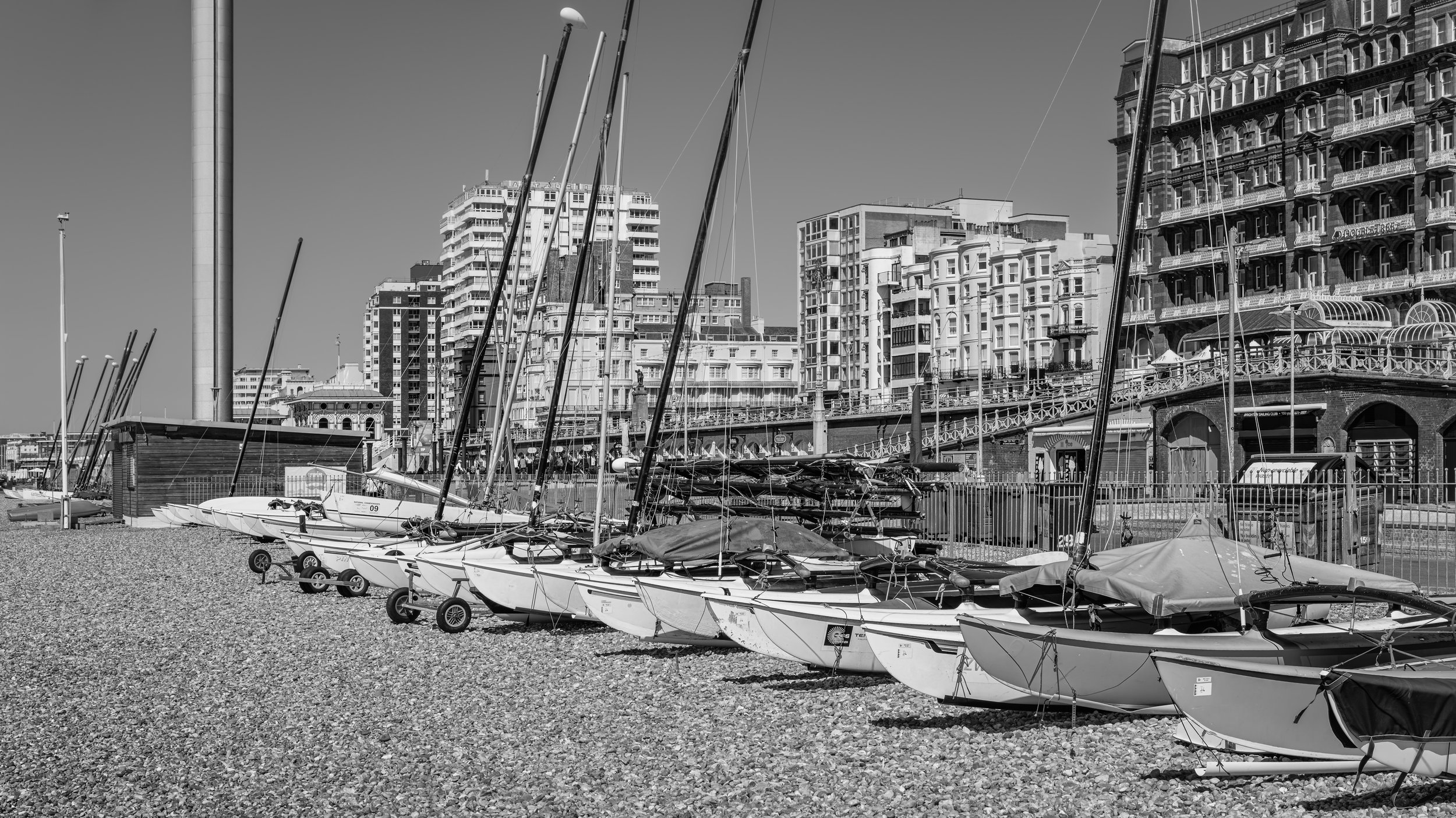 Sailboats docked on a pebble beach with tall buildings and city infrastructure in the background, black and white photo.