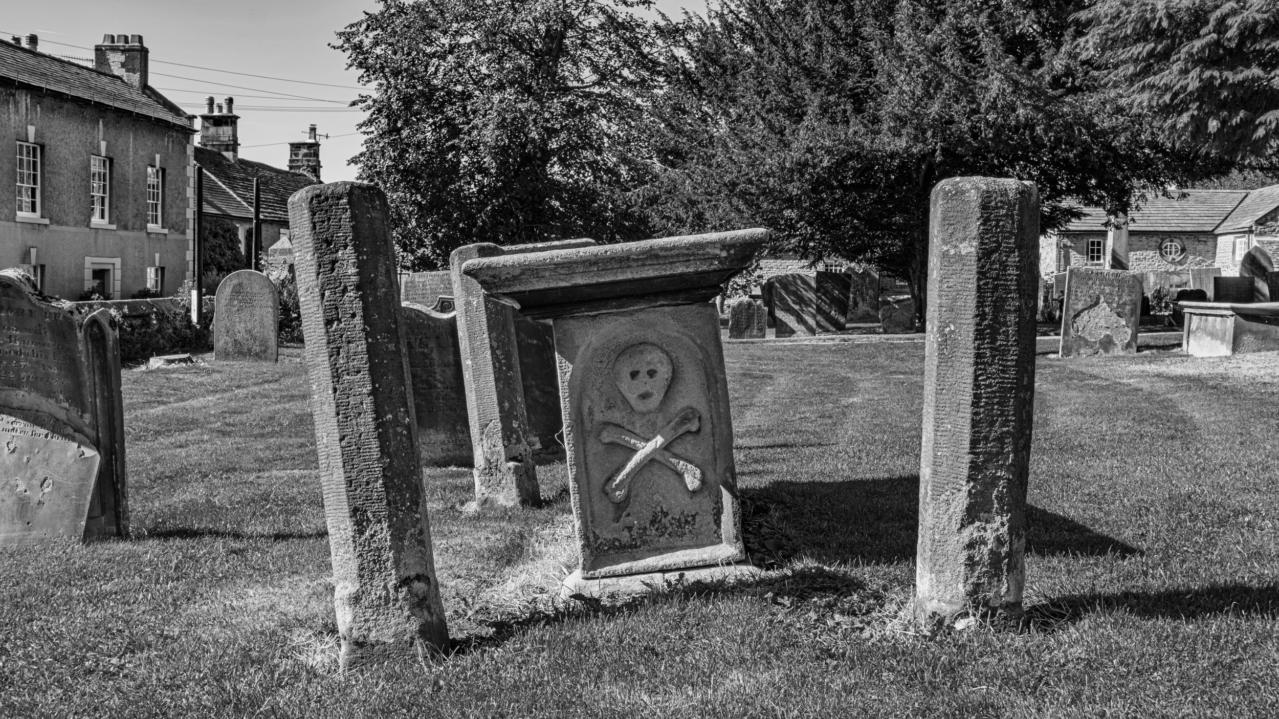 Black and white photo of a graveyard with old and weathered tombstones, some featuring decorative carvings and symbols, surrounded by grass and trees, with a row of houses and utility wires in the background.