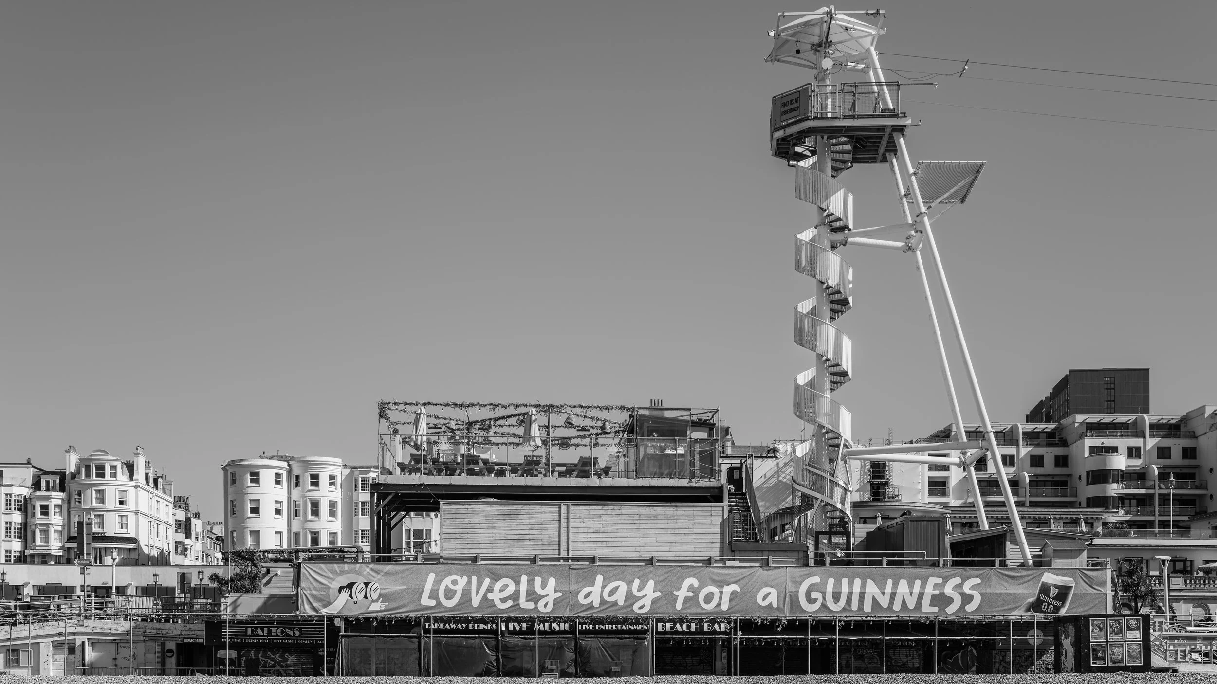 A panoramic view of a cityscape featuring a large Ferris wheel with a spiral staircase, buildings with various architectural styles, and a banner advertising a Guinness event, with a clear sky in the background.