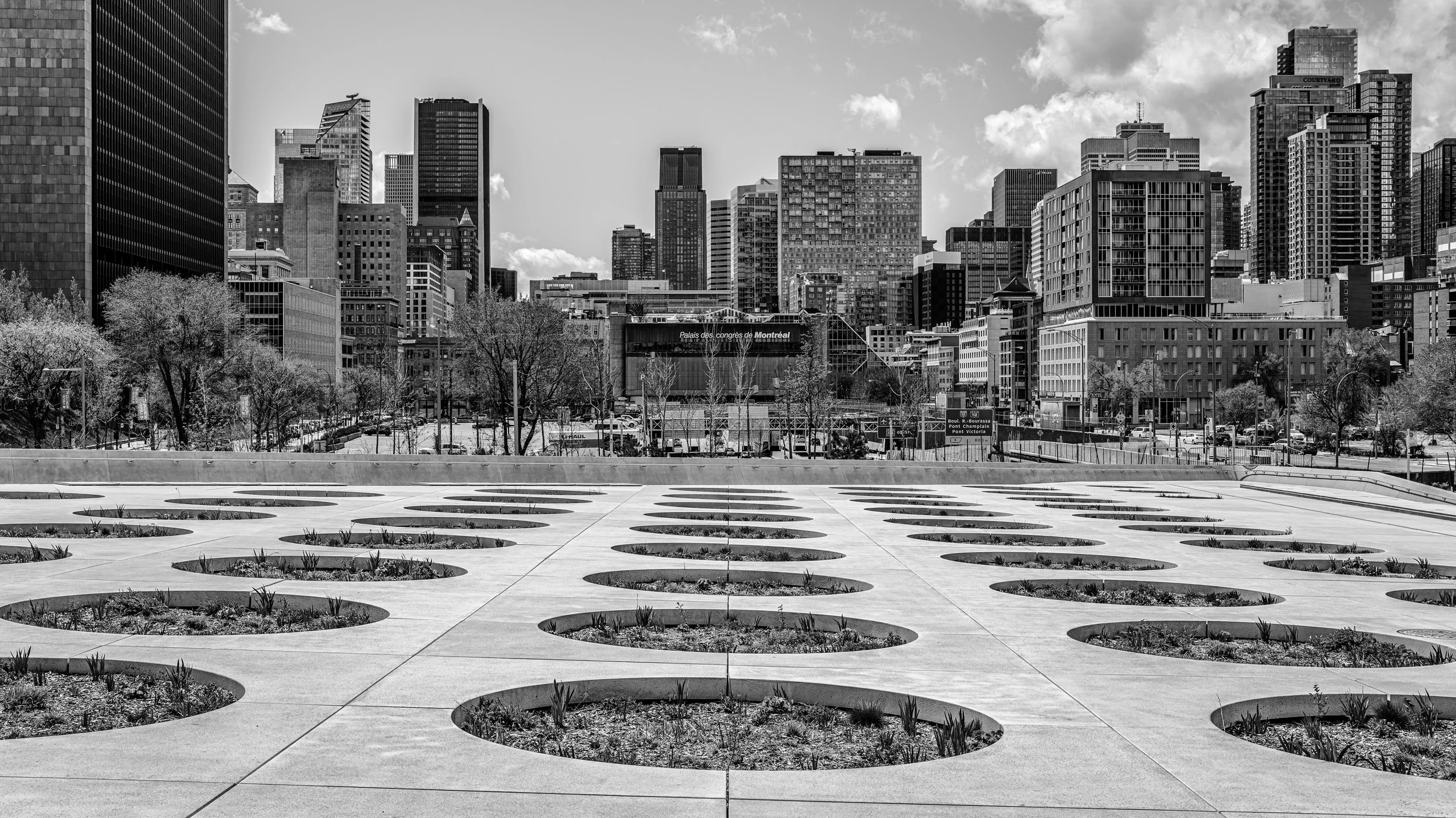 A black and white cityscape of Montreal with modern skyscrapers and trees in the foreground, including a sign indicating 'Palais des congrès de Montréal'.