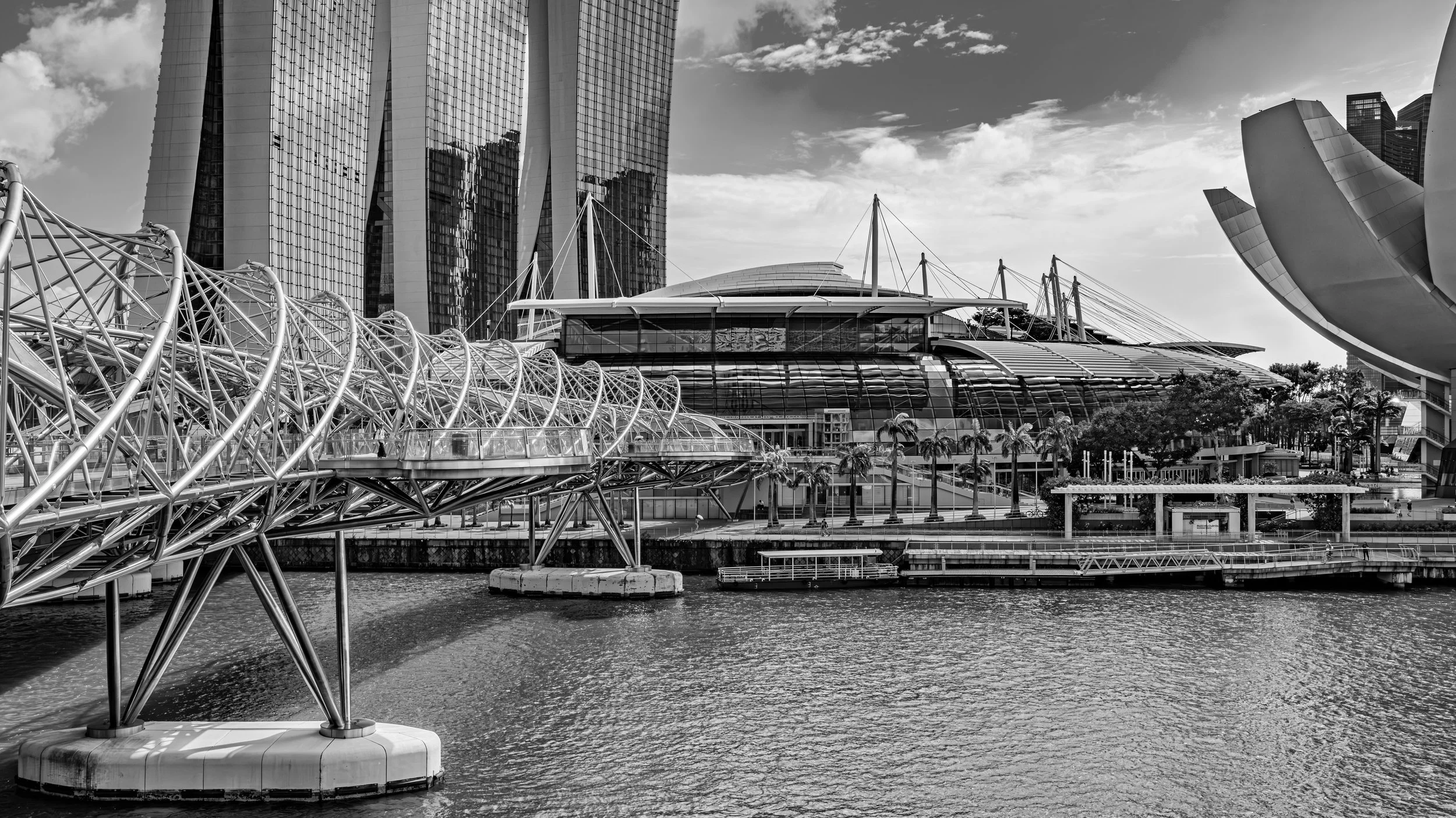 Black and white photo of downtown Singapore skyline with modern buildings, the Esplanade theatre, and the Helix bridge over the water.
