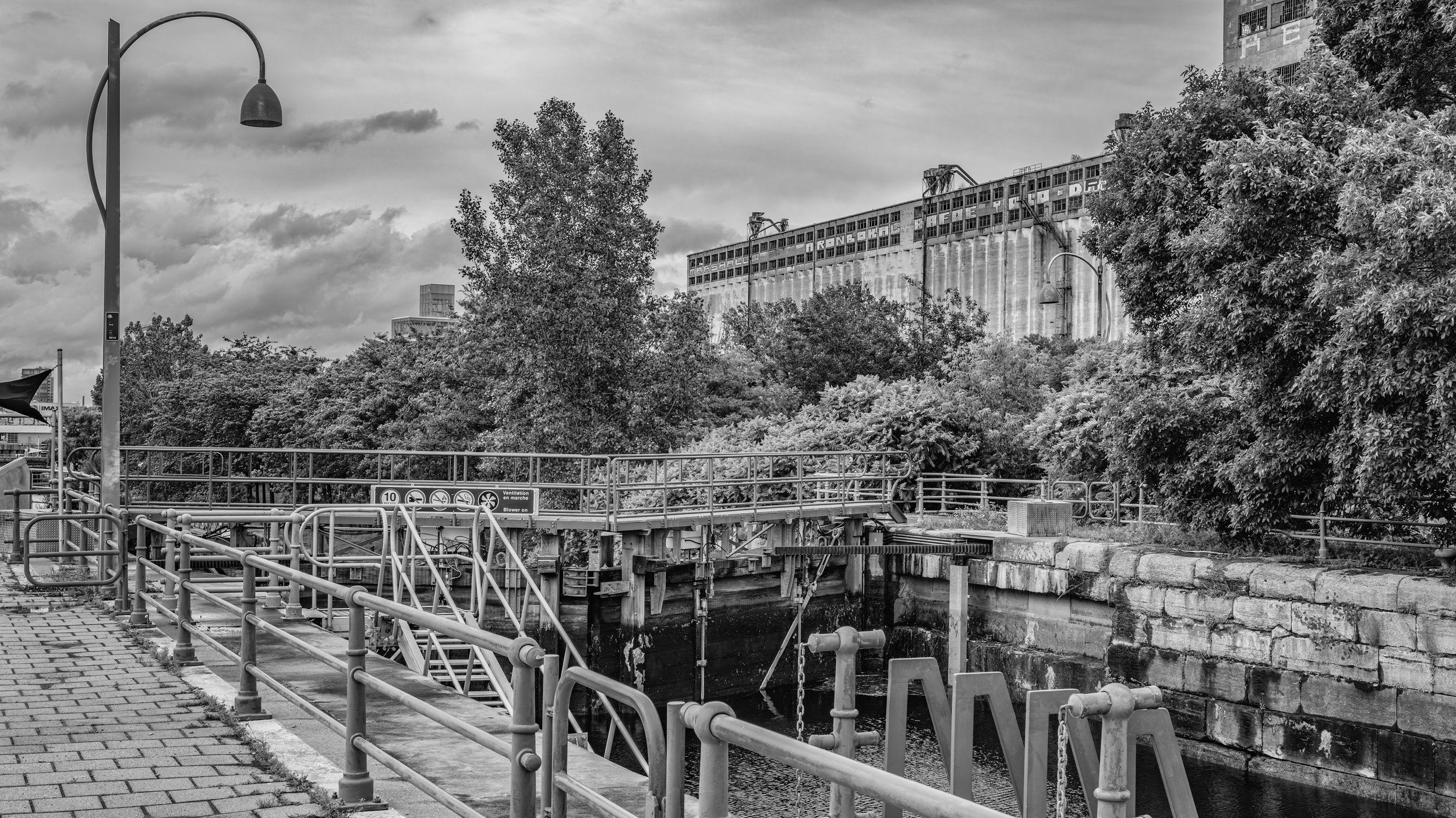 Black and white photo of a cityscape featuring a large, abandoned building with graffiti, trees, street lamps, and a canal or waterway with metal railings and a dock in the foreground.