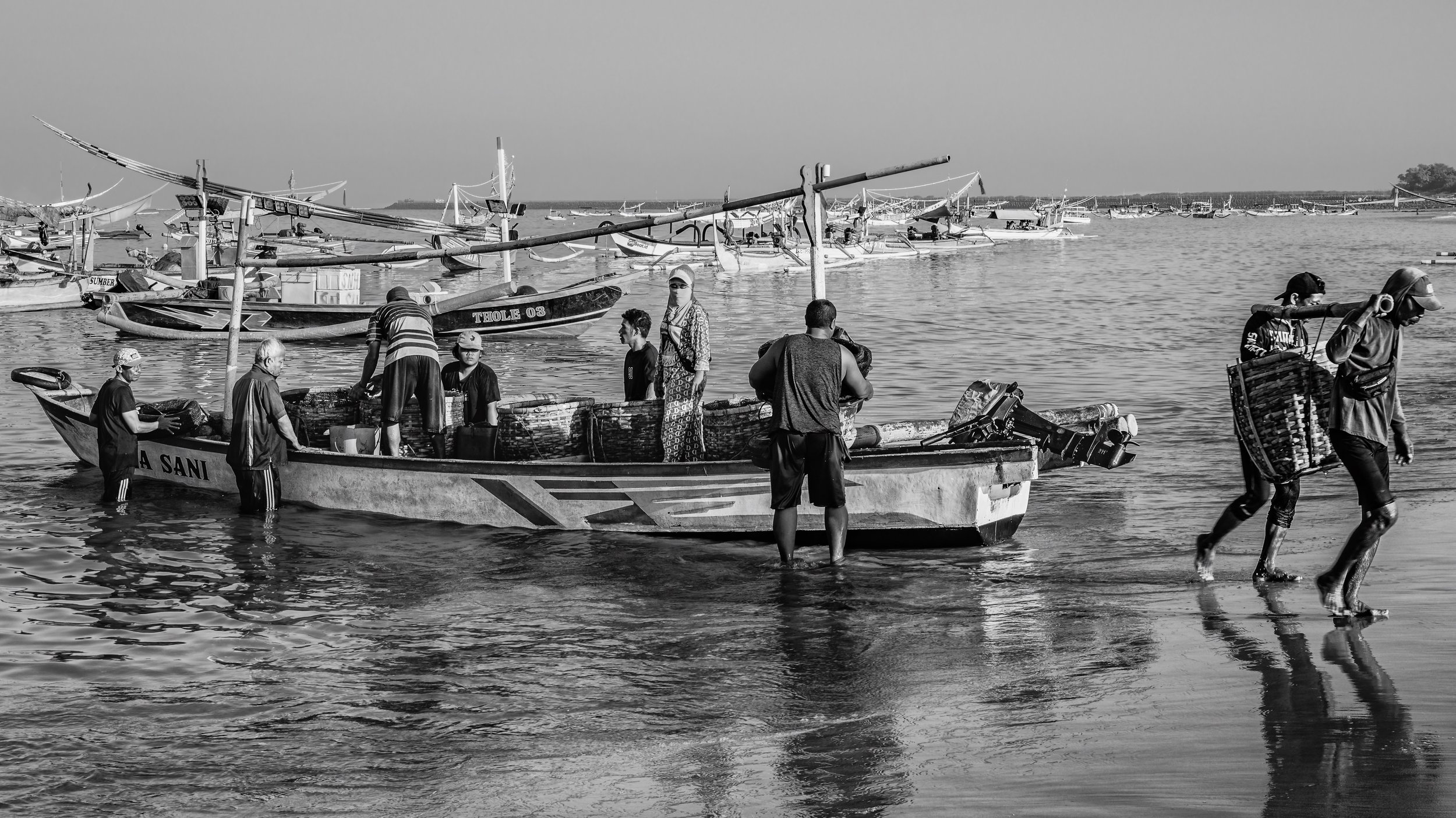 People loading boats at a harbor with numerous boats anchored in the water and land in the background.