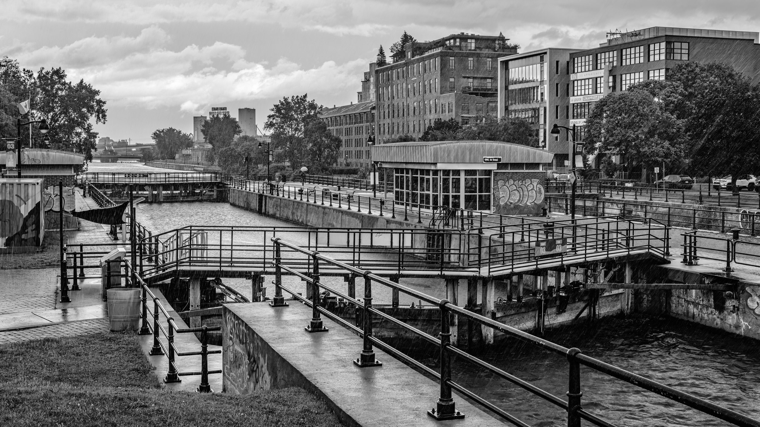 Black and white photo of an urban canal with a walkway, benches, and railings, surrounded by modern apartment buildings, trees, and cloudy sky.