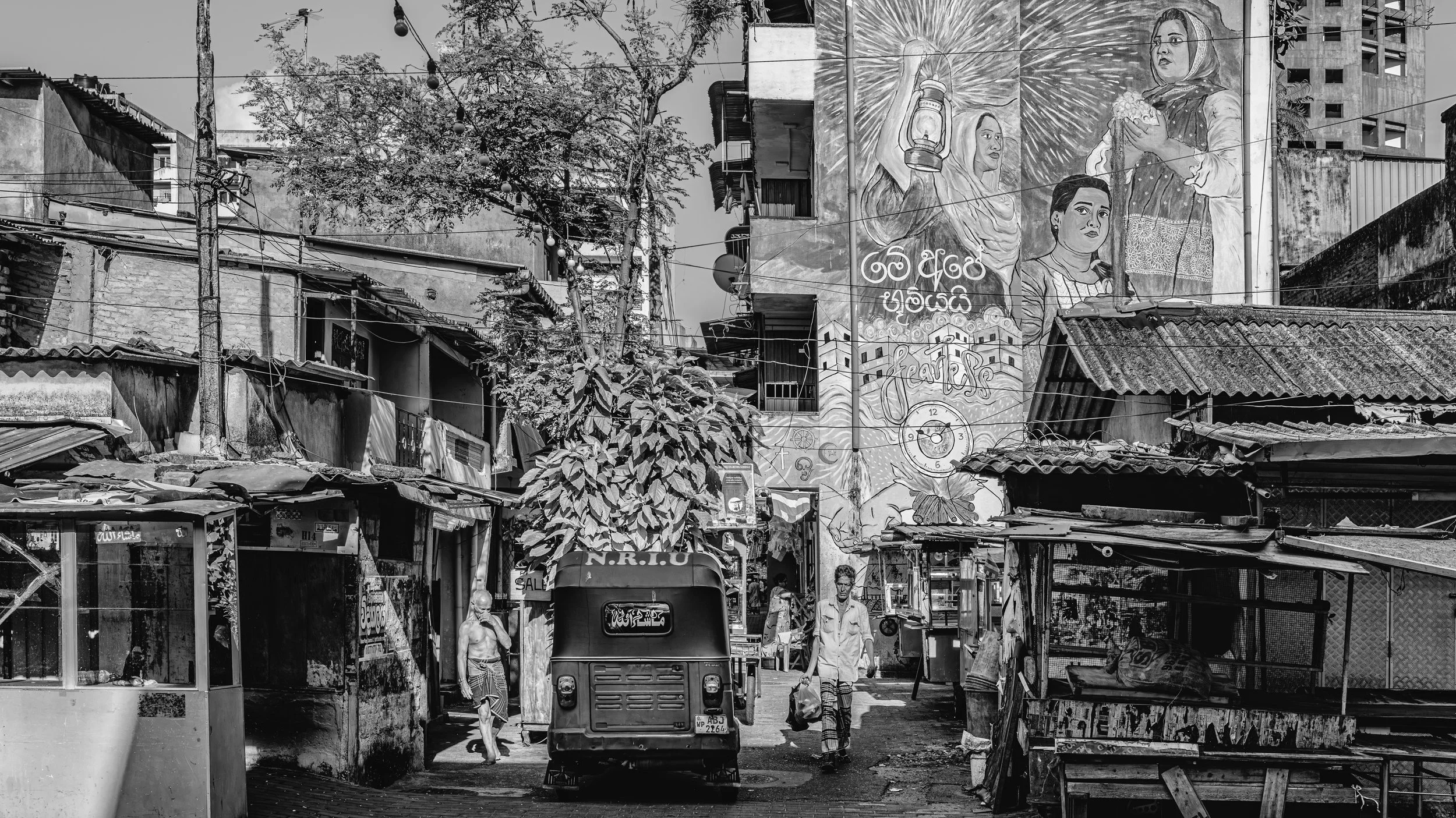 A black-and-white street scene in a neighborhood with small shops and makeshift structures. People walk on the street, and there is a tuk-tuk vehicle in the center. The background features a large mural with images of women, a lantern, and text in a 