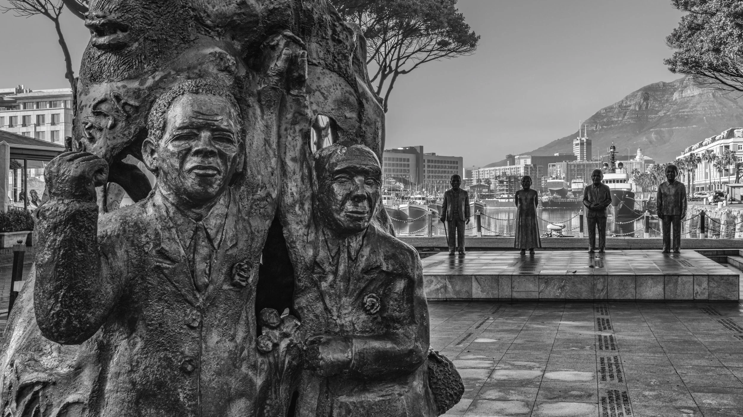 A black and white photo of a statue with several human figures behind it, set against a marina with boats and a cityscape, and a mountain in the background. Cape Town, South Africa.