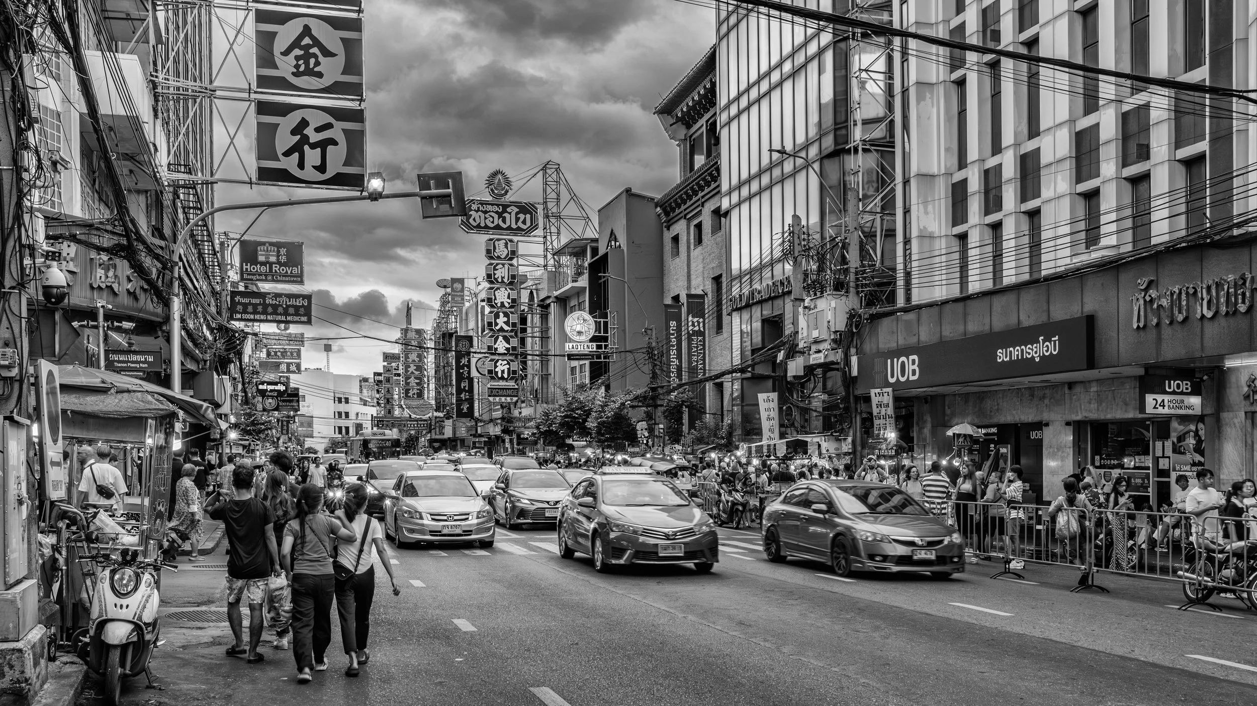 A busy city street filled with cars, pedestrians, and tall buildings with numerous signs in Asian script, including a bank and hotel, under a cloudy sky. Bangkok. Thailand.
