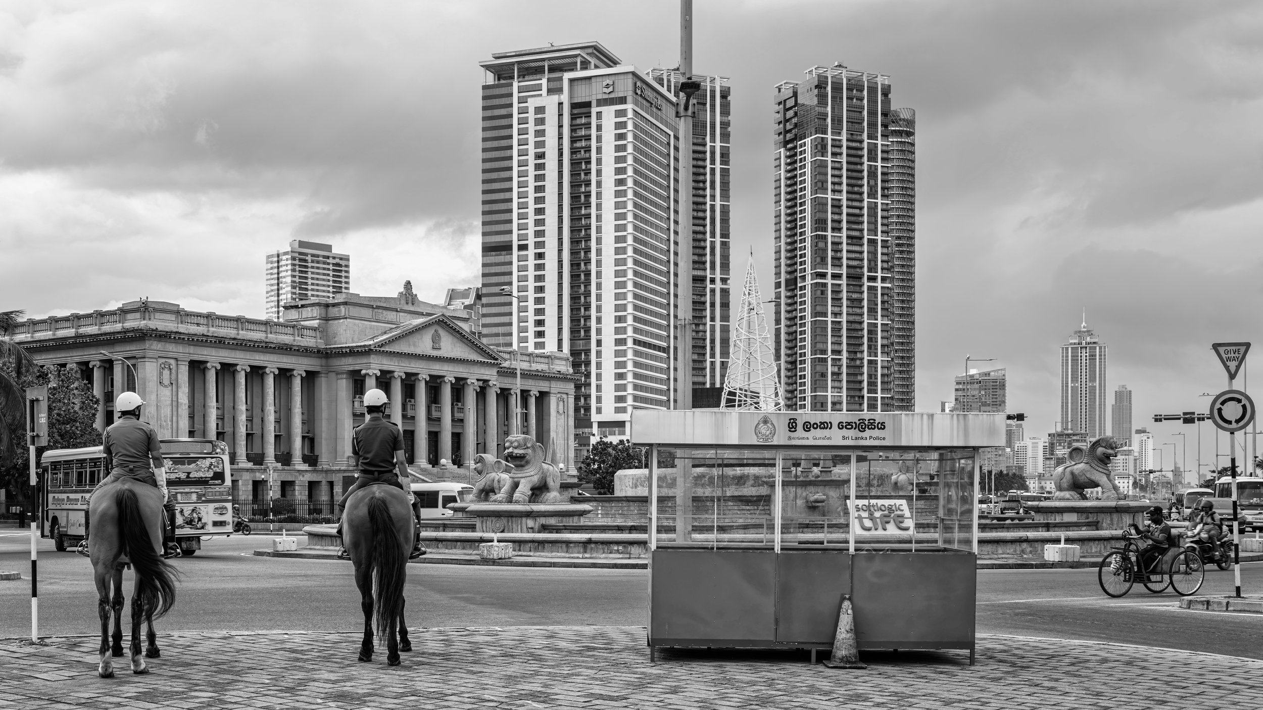 Black and white photo of a city street scene with modern tall buildings, Sri Lanka Police signage, two police officers on horses, a lion statue, and a few people riding a bicycle and motorcycle.