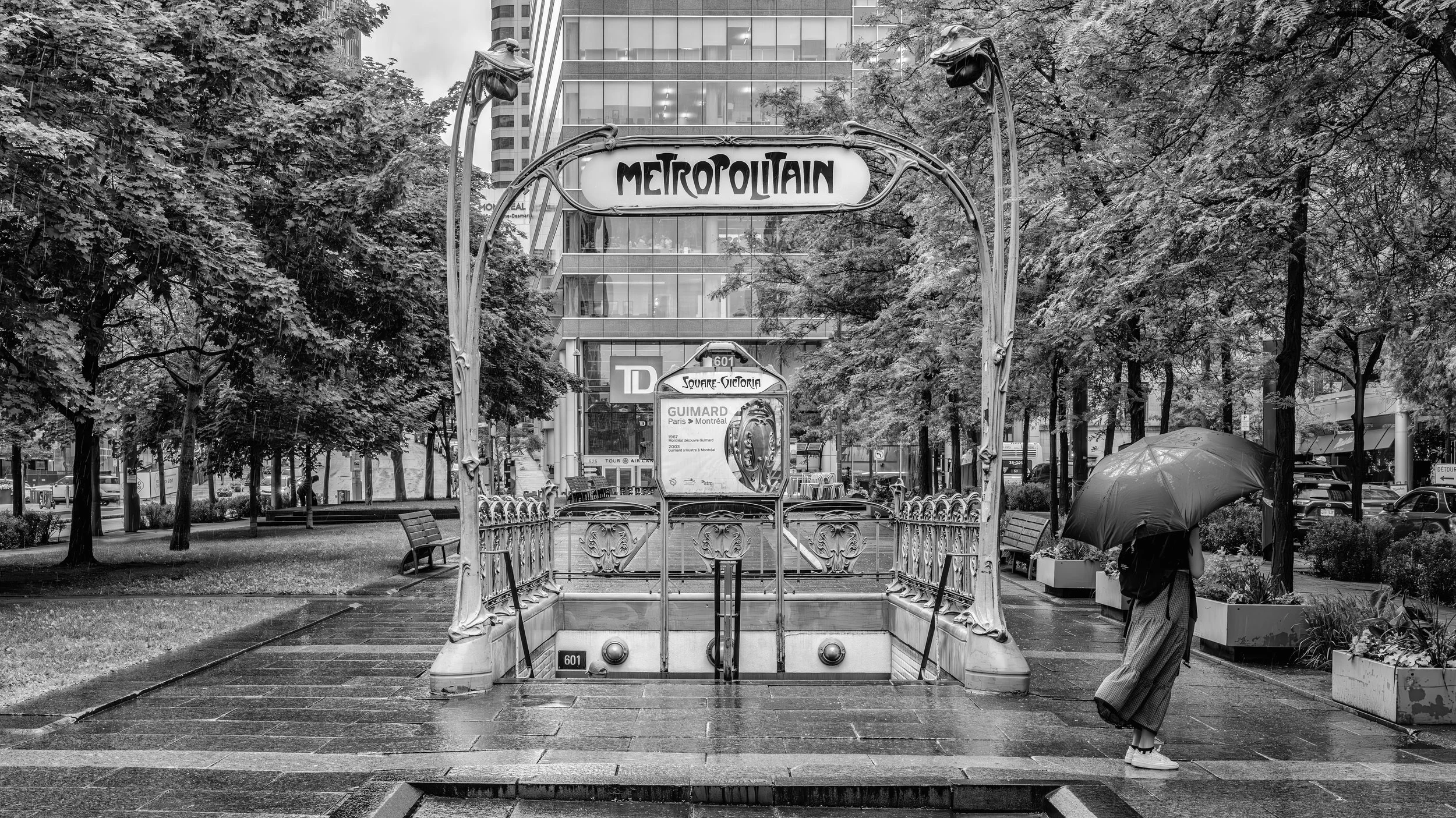 A black and white photo of the entrance to the Metro Metropolitain subway station in an urban area with tall buildings. The station sign is ornate, with decorative metalwork, and the word "METROPOLITAIN" displayed in bold letters. There are trees, be