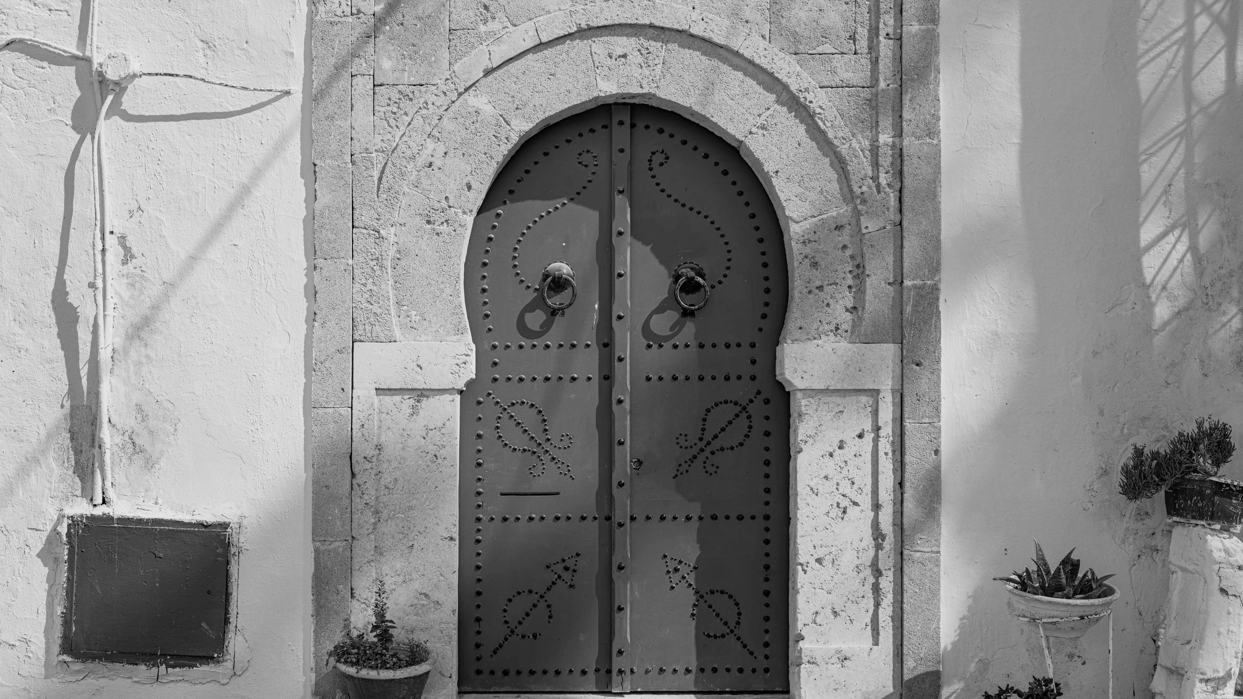 Black and white photo of an arched wooden door with decorative ironwork and large round handles, set into a stone wall. There are potted plants and shadow patterns on the wall surrounding the door.