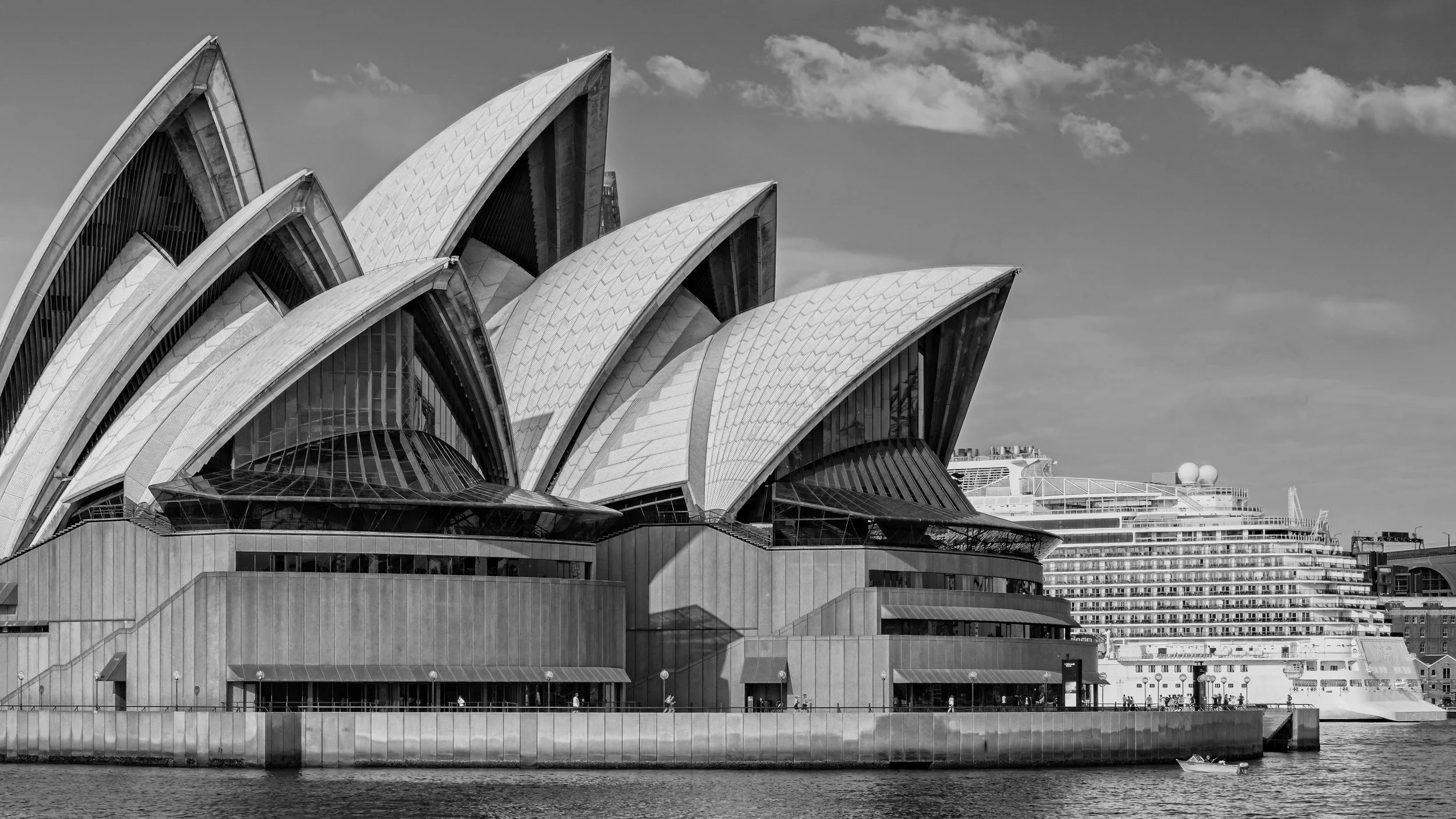 A black and white photo of the Sydney Opera House with a cruise ship docked nearby and a small motor boat on the river in the foreground. Sydney, New South Wales, Australia.