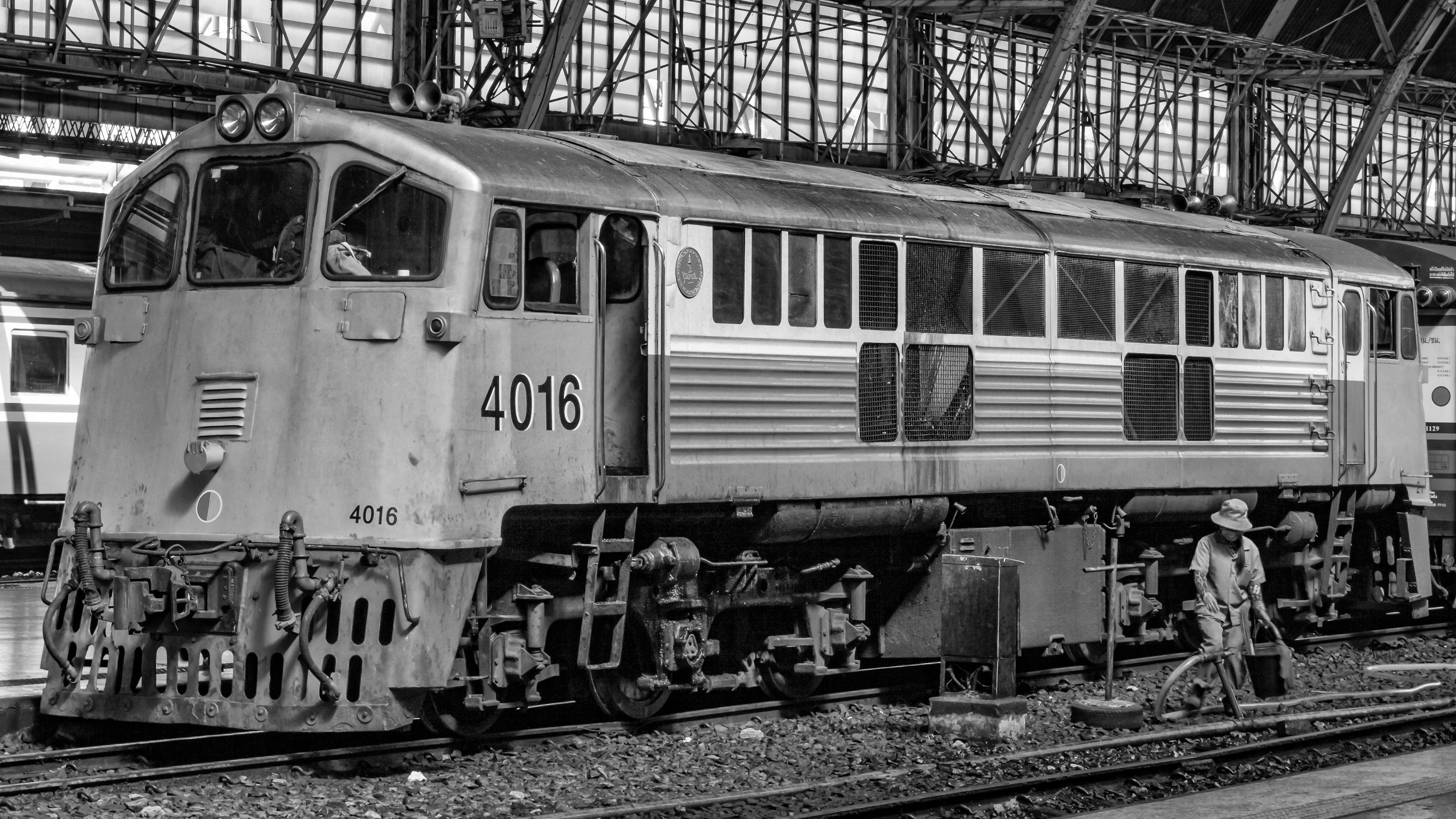 A black and white photograph of an old locomotive train at a train station. The number 4016 is visible on the front and side of the locomotive. There is a person wearing a hat working nearby, and the station structure with metal beams and glass ceili