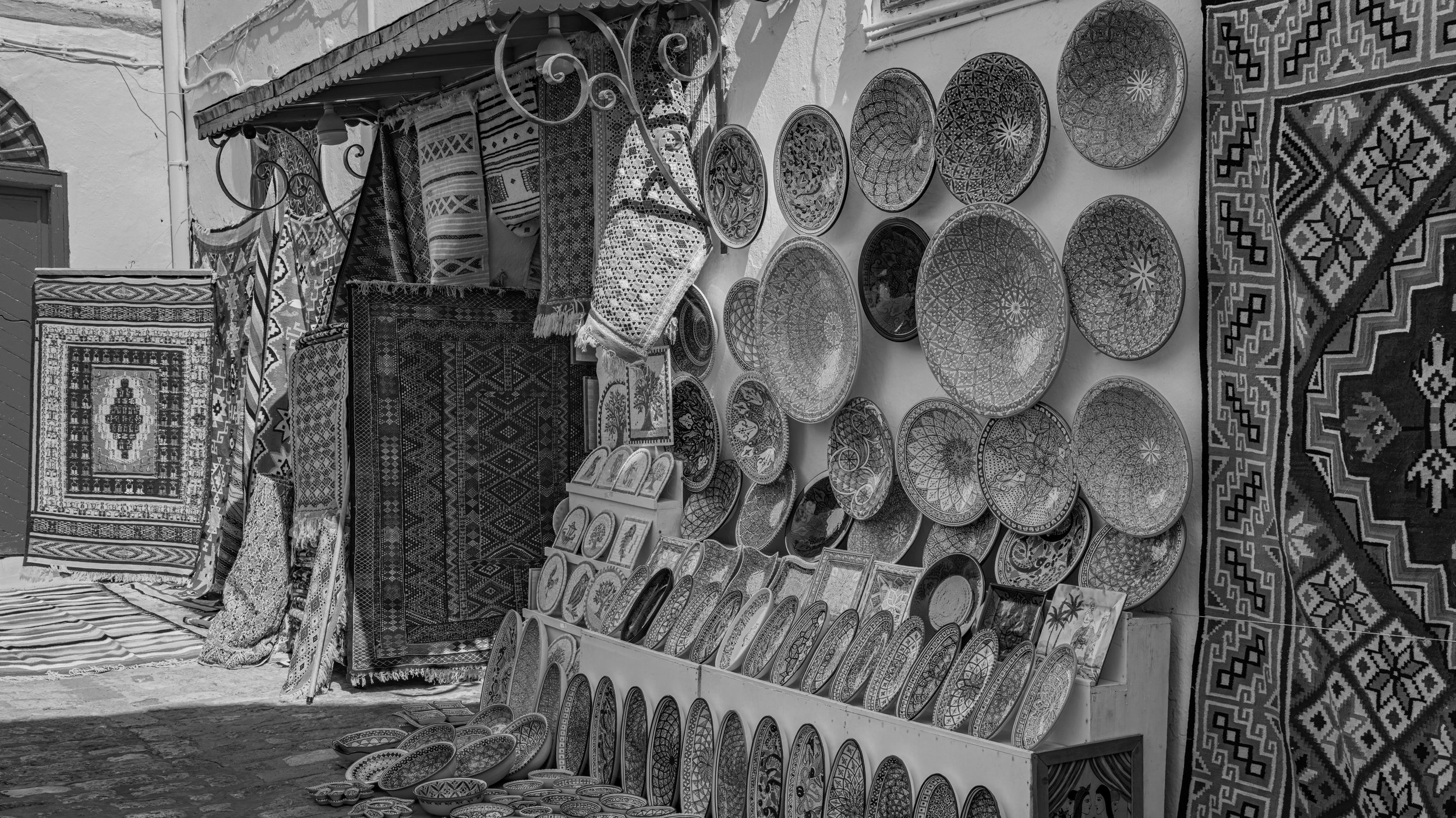 Display of various patterned woven rugs and plates with intricate geometric designs at an outdoor market.