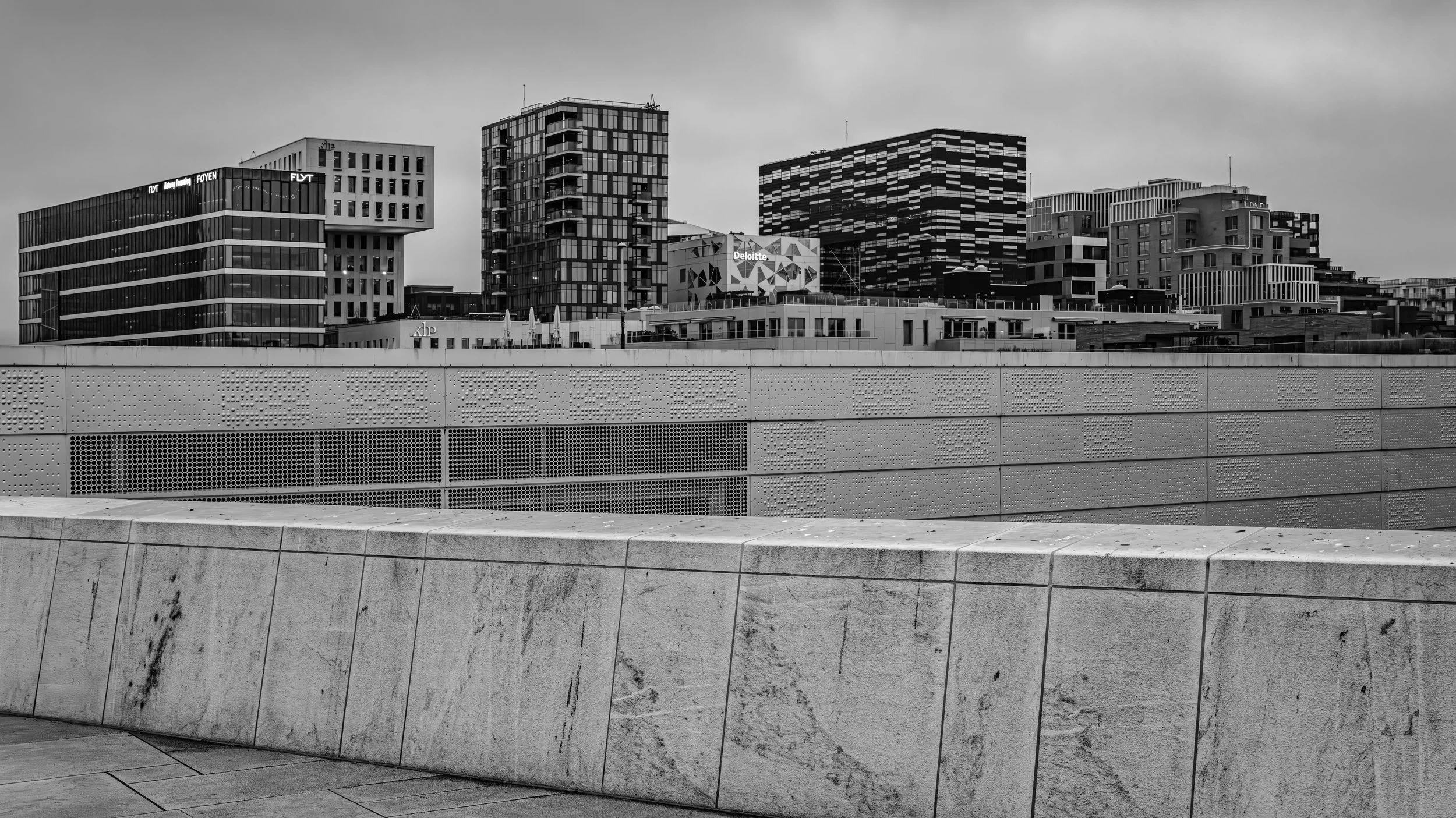 A cityscape with modern high-rise buildings, including office buildings and apartments, in black and white, with a textured concrete structure in the foreground.