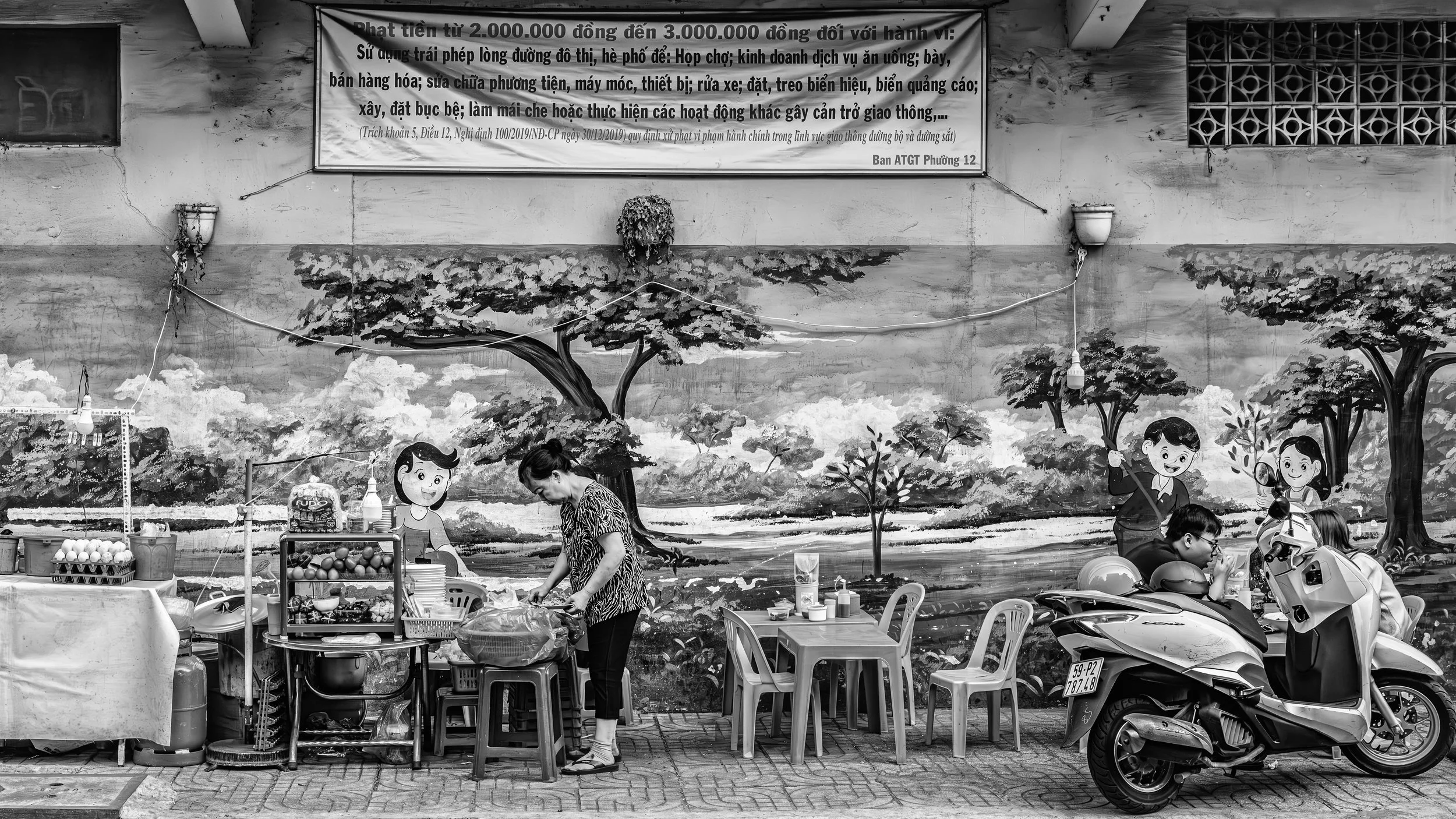 A street scene with a mural painting of trees and children playing. A woman is working at a food stall with a table of eggs and other items, while scooters are parked in front.
