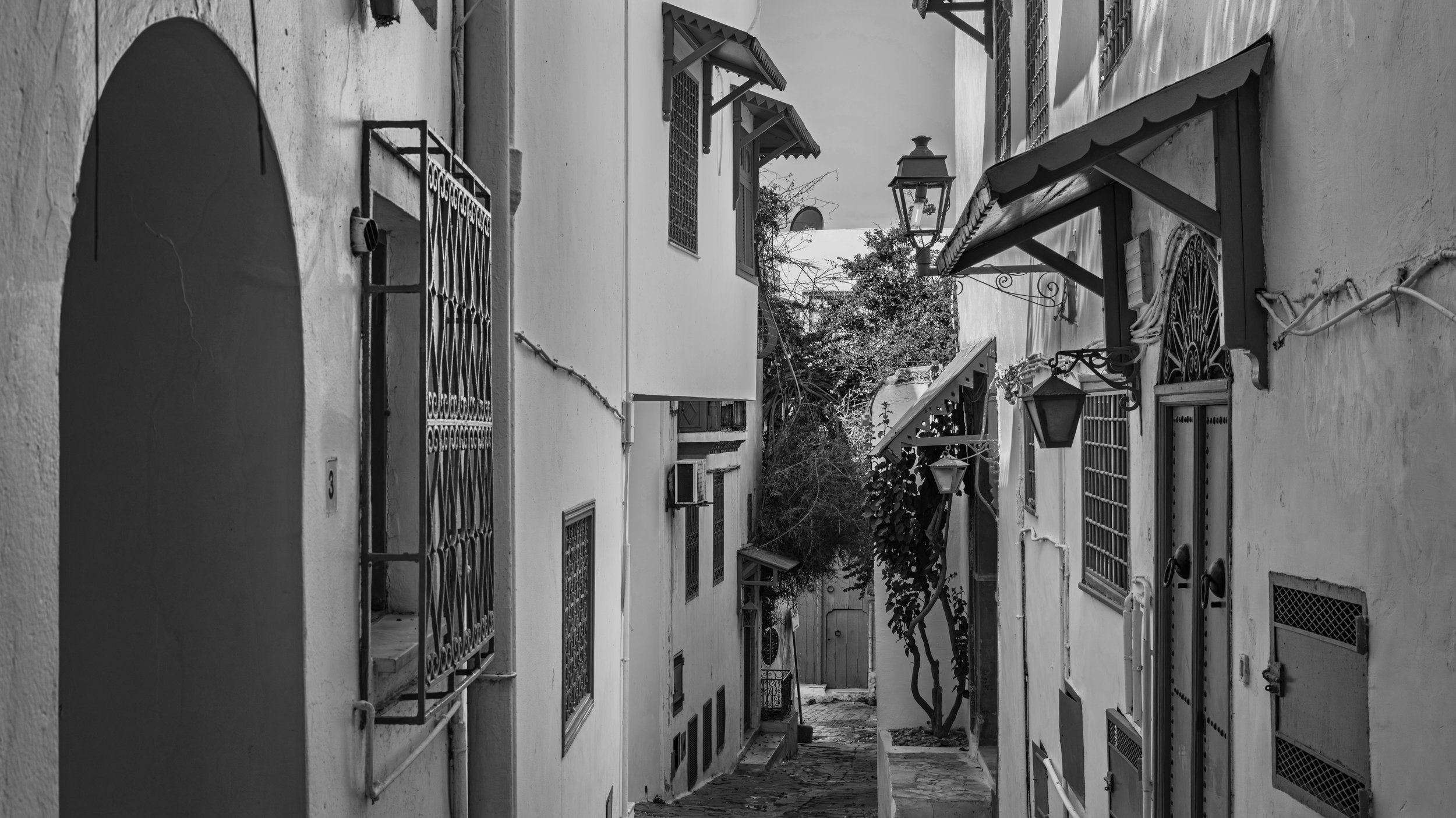 A narrow alleyway between white buildings with barred windows, street lamps, and rooftops with awnings, in black and white.
