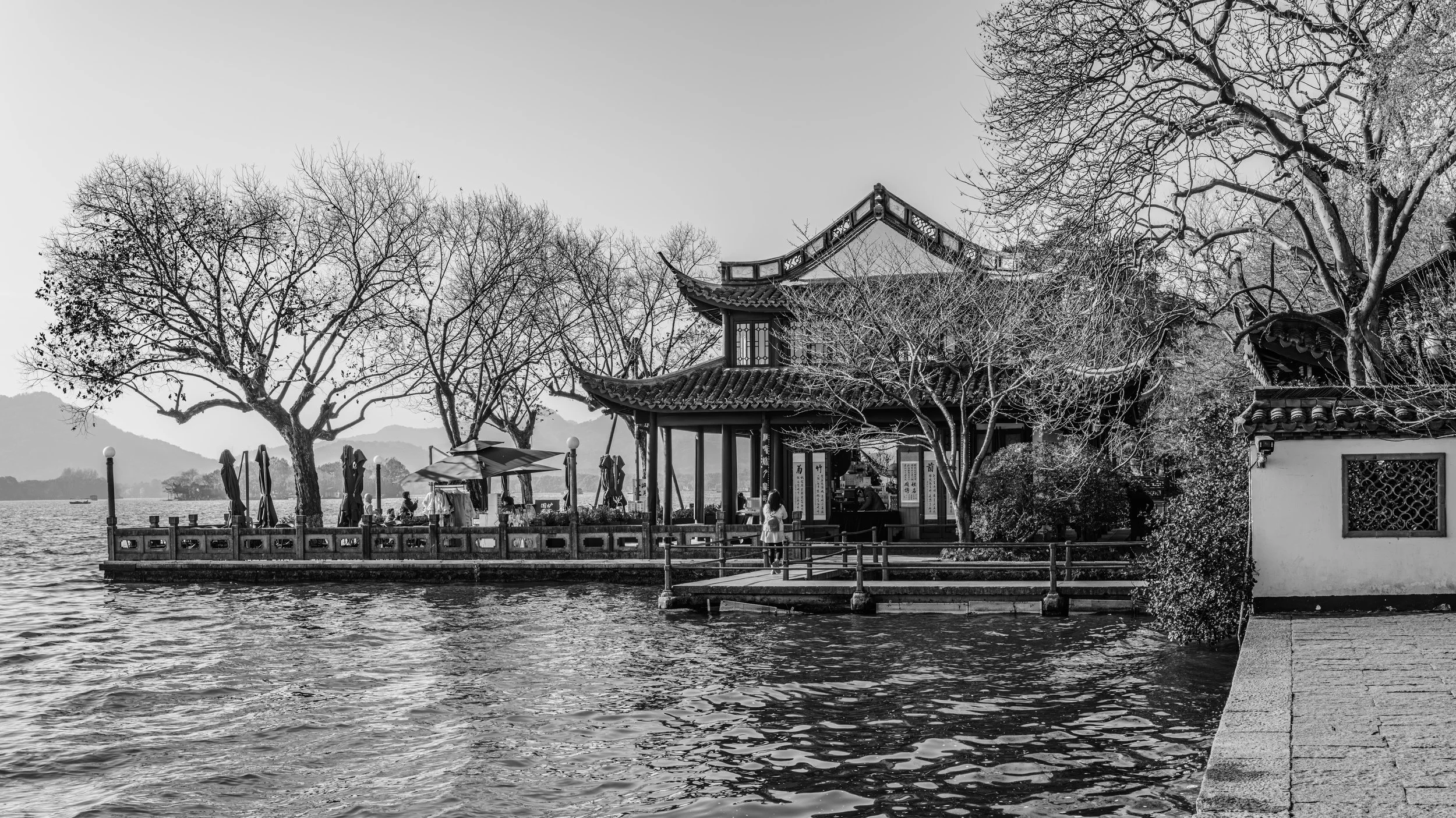 A lakeside scene in black and white featuring traditional Asian-style architecture with a curved roof, trees without leaves, and a deck with umbrellas and people by the water.