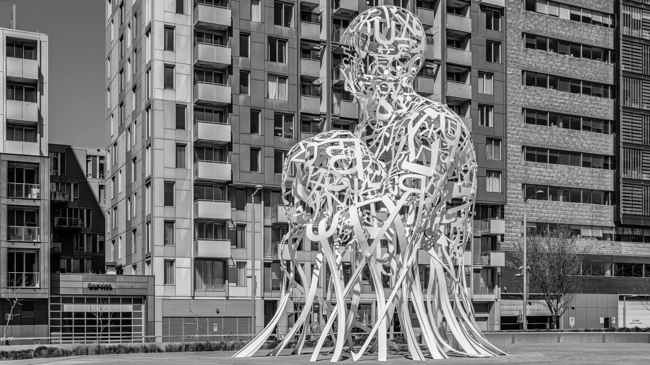 Street art sculpture resembling a human head and shoulders made of intertwined white strips, with a background of modern high-rise apartment buildings.