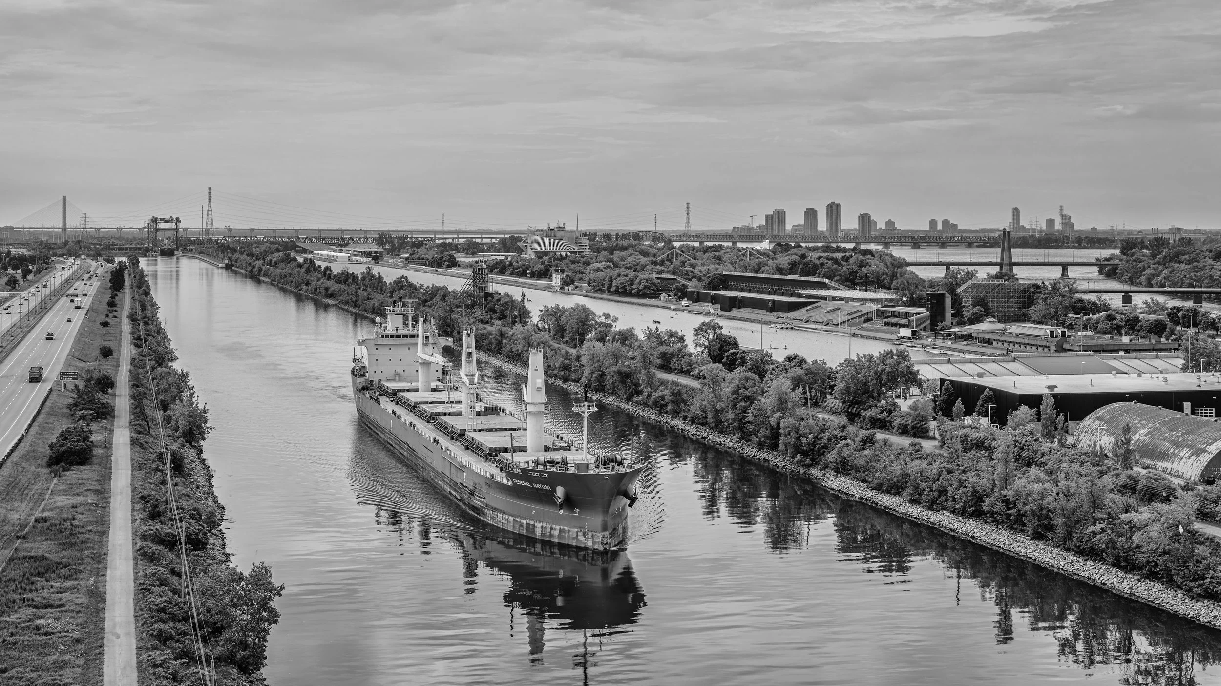 A black and white photograph of a cityscape with a river in the foreground. A large cargo ship is sailing on the river, and there are roads with vehicles on the left side. The background shows a skyline with tall buildings, bridges, and a partly clou
