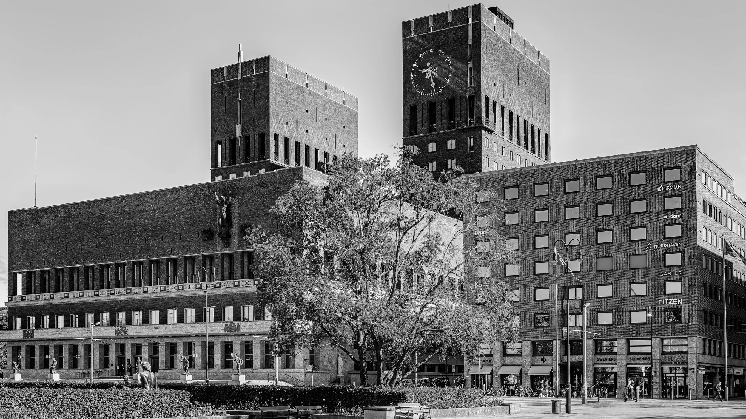 A black and white photo of a cityscape featuring a large brick building with two towers, one of which has a clock. There are trees, benches, and people walking in front of the building.