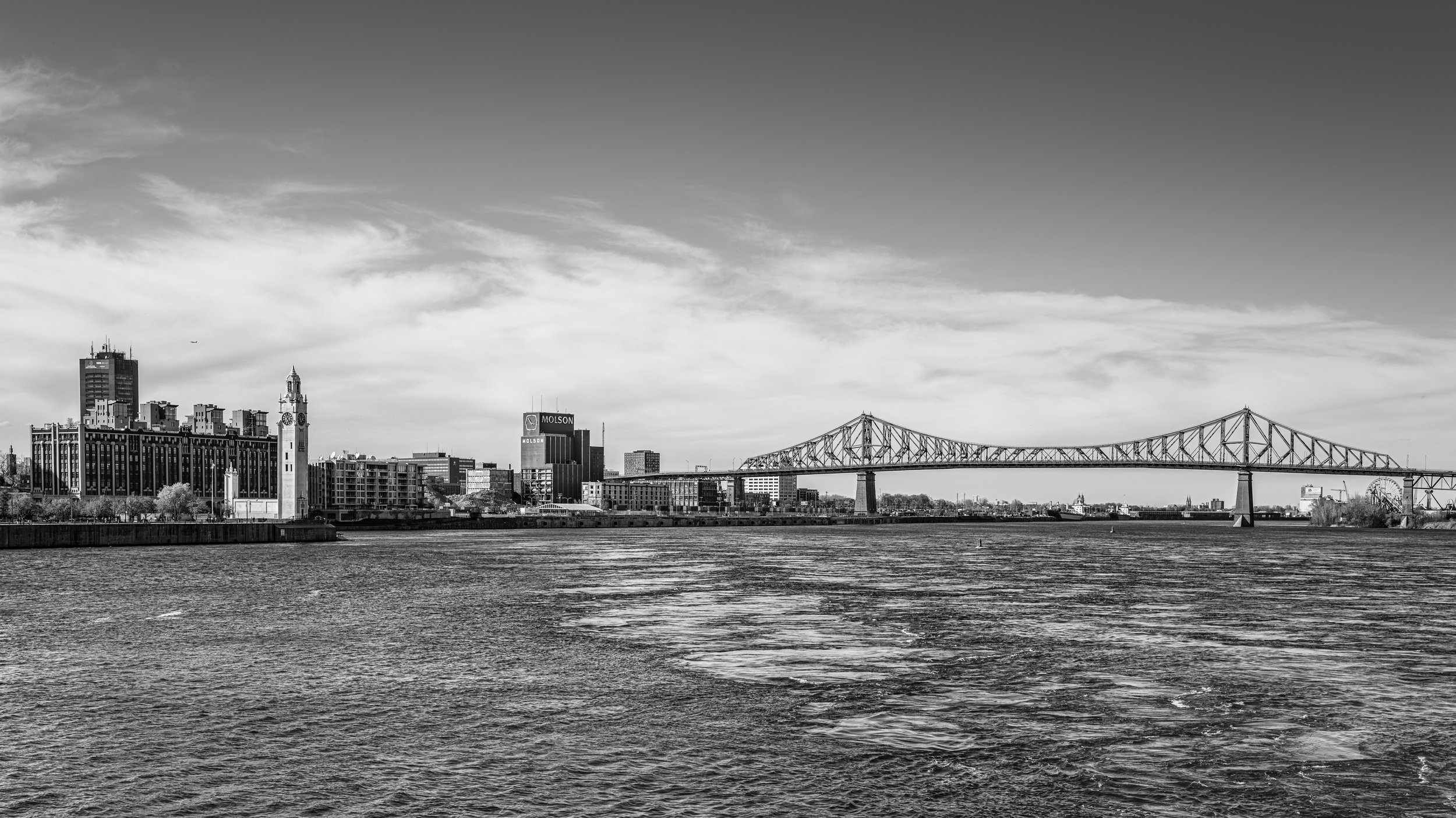 Black and white photograph of a city skyline with a bridge over water, featuring tall buildings, a clock tower, and a Ferris wheel in the background.