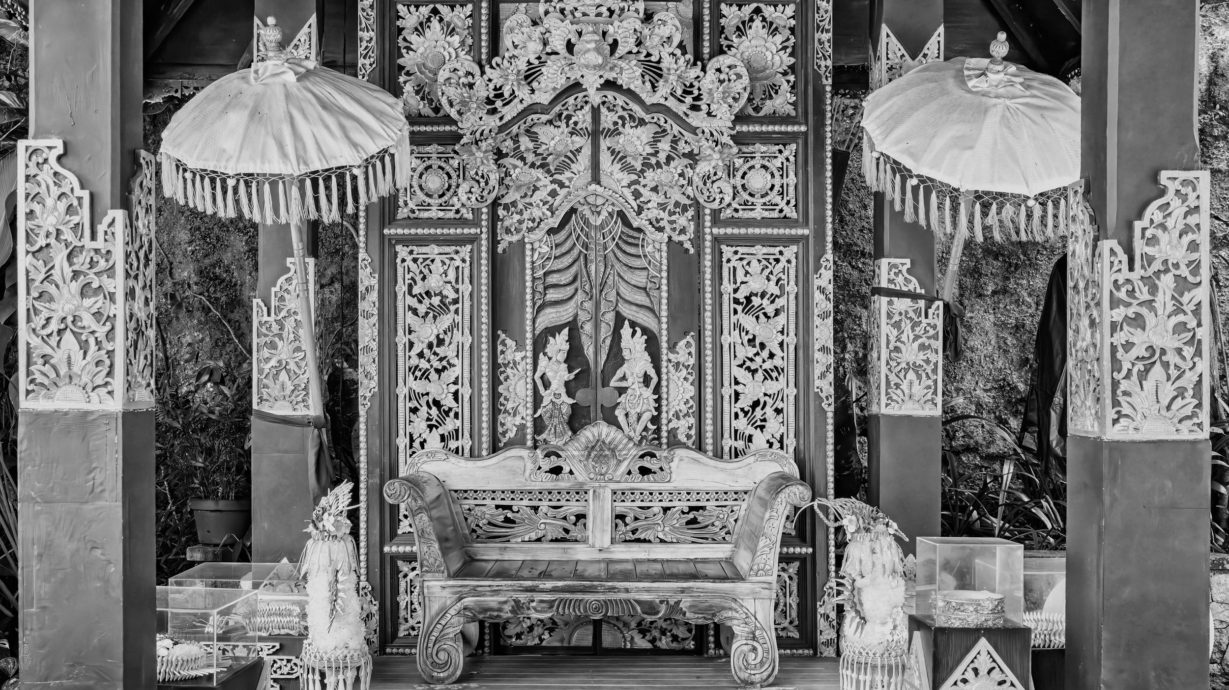 Elaborately carved wooden door flanked by two umbrellas with tassels, set in a decorative frame, with a small bench in front and plants on either side.