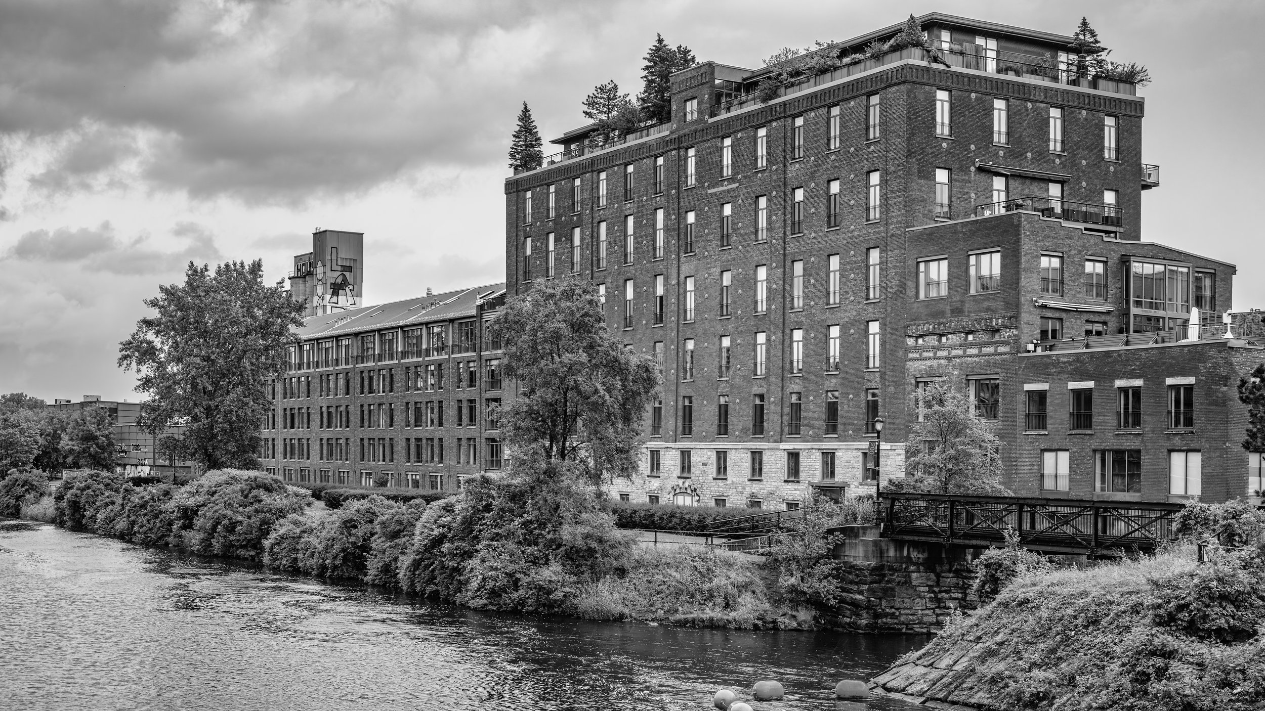 Black and white photo of multi-story brick buildings next to a river, with trees and a cloudy sky in the background.