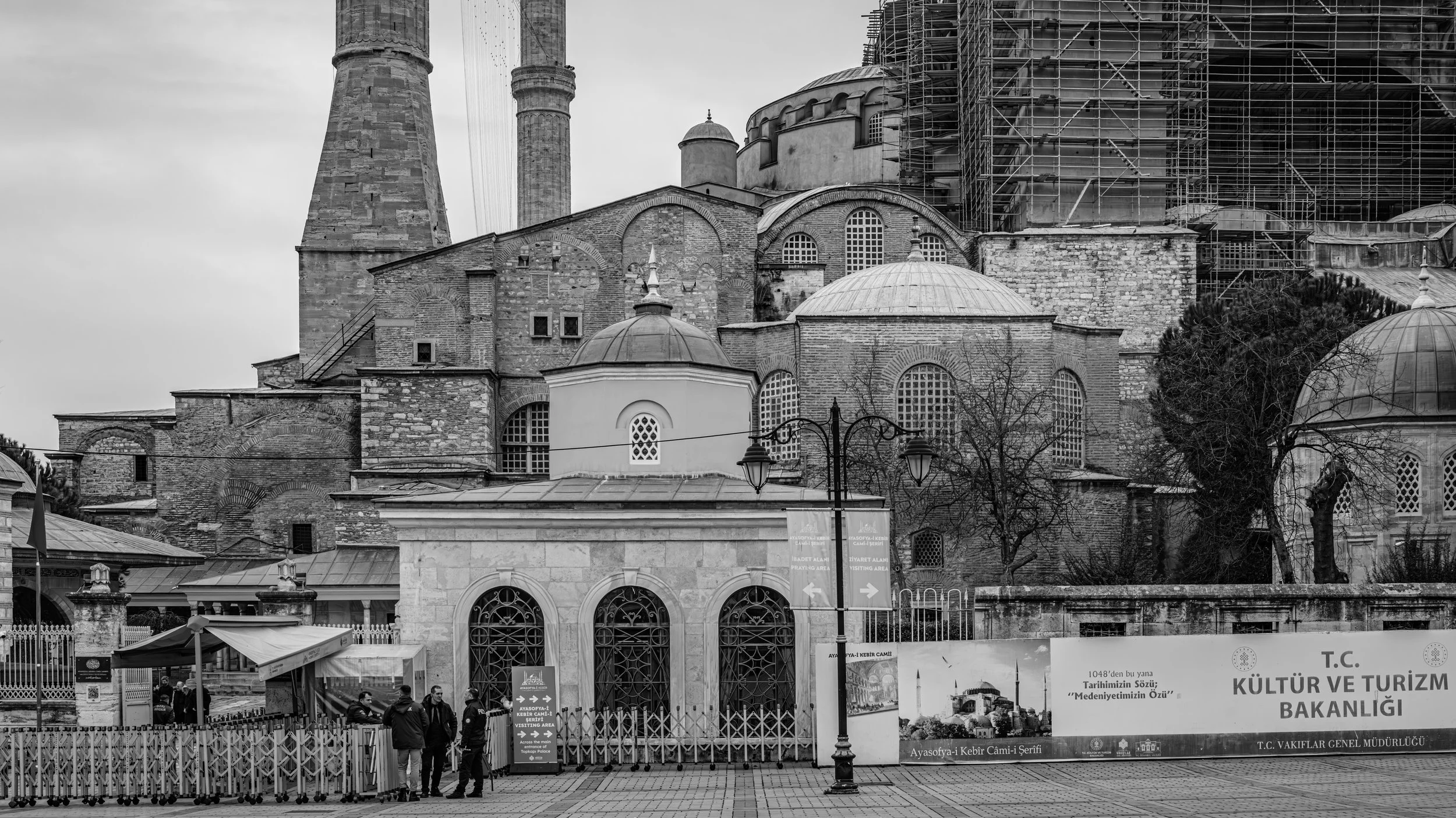 Black and white photo of an old mosque with domes and minarets, partly covered in scaffolding, with leafless trees and street lamps in front, and a group of people standing near the entrance.